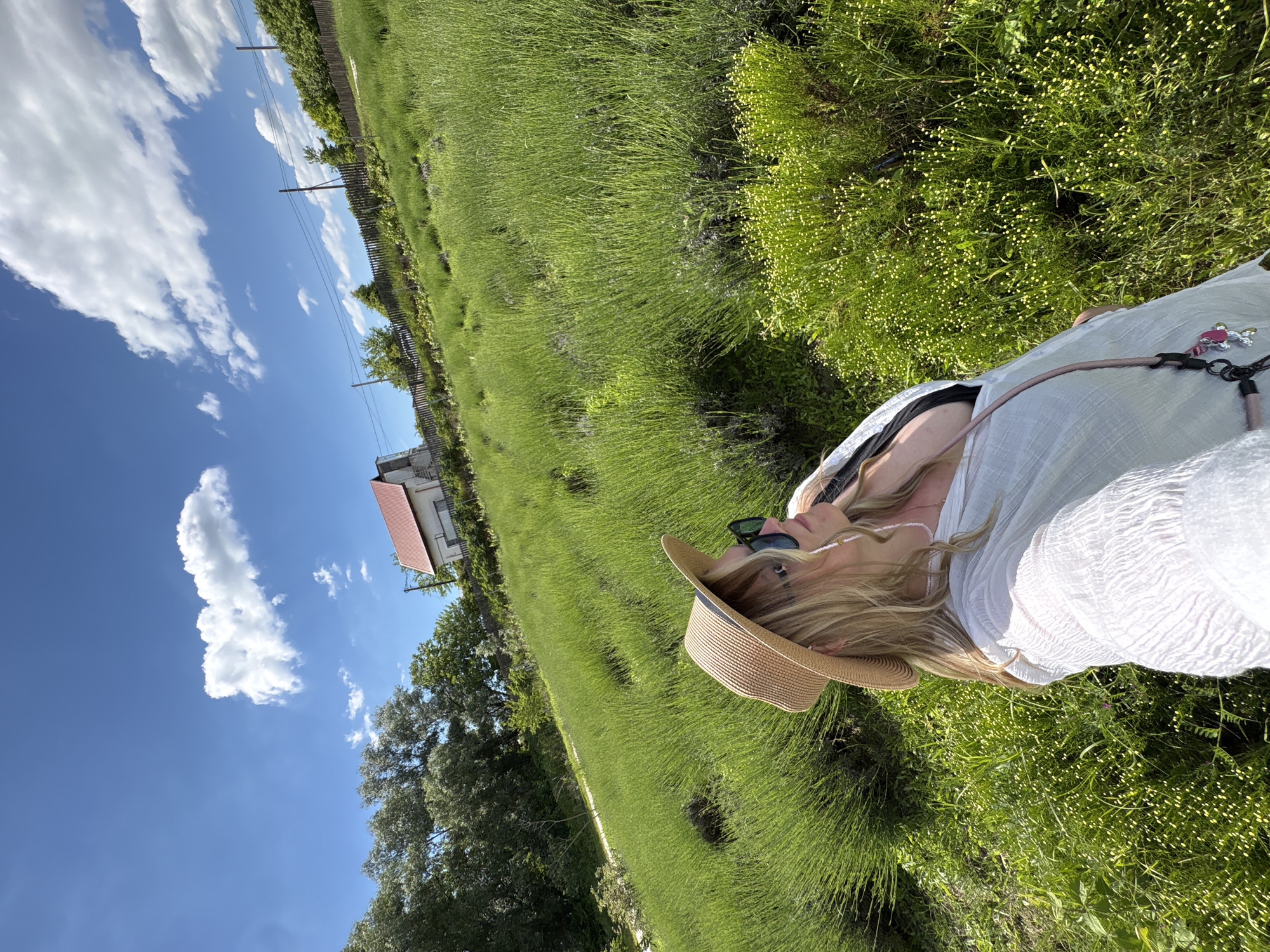 This media file captures an outdoor scene in Gay-Kodzor, Russia, on a bright, sunny day. The image is composed with a wide expanse of vibrant green vegetation dominating the lower and central parts of the frame, possibly a field of young crops like lavender. The sky is a clear, brilliant blue with scattered white, fluffy clouds, indicative of good weather.
A person is visible on the right side of the frame, from the chest up, with their head tilted slightly upwards. They have long, blonde hair and are wearing a light-colored, possibly white, textured top or dress. A wide-brimmed straw hat covers part of their head, and sunglasses are worn either on their face or perched on their head. A necklace with small white beads is visible around their neck, and a strap, possibly from a bag or camera, with pink and silver charms, crosses their chest.
In the mid-ground and background, on the left side, there are several large green trees and a visible wooden fence line. Further back, a house with a distinct red-tiled roof can be seen amidst more greenery. Utility poles with overhead wires are also present, running across the landscape. The overall atmosphere suggests a peaceful, leisurely moment spent outdoors in a rural or semi-rural setting, enjoying the sunshine. There is no visible text in the image.