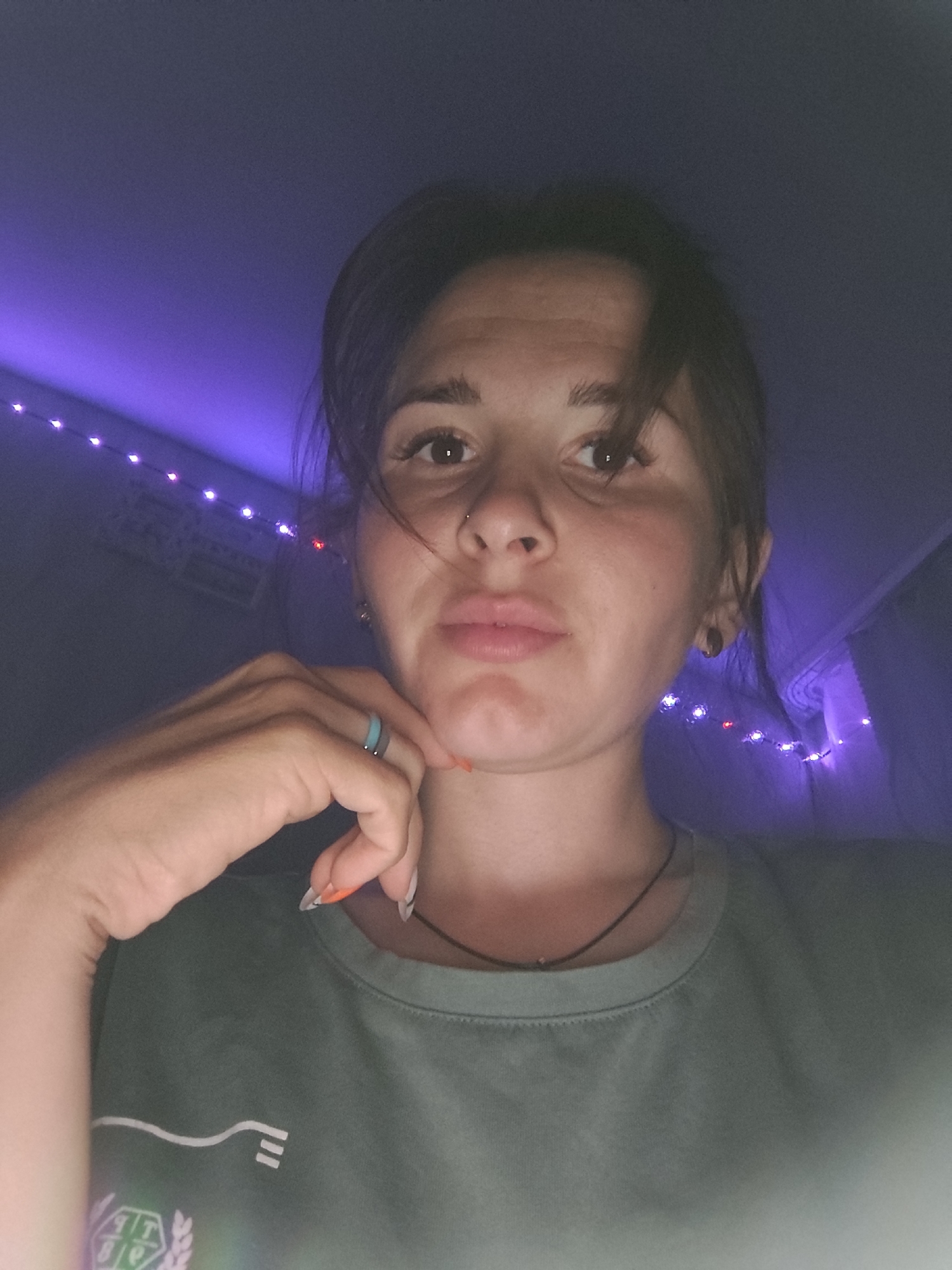 A single female subject is depicted indoors in a dimly lit environment, confirmed to be in the city of Фоки, Russia. The subject has dark hair, brown eyes, and a light to medium skin tone. A small nose piercing is visible on her right nostril, and she wears small, dark earrings in both ears. Her lips are slightly pursed. She is wearing a muted green/teal crew-neck t-shirt with a white wavy line design and a partial emblem visible on the lower left front. A thin, dark necklace with a small pendant is around her neck. Her left hand is raised, with fingers resting beneath her chin. The fingernails on her left hand feature a French tip design, with an additional orange accent on the ring finger. A light blue and grey ring is worn on her left ring finger. The background is dark, illuminated by purple ambient lighting that casts a glow on a ceiling or wall. A string of small, round LED lights, primarily purple with a few red lights, is visible horizontally across the background. The overall lighting suggests an evening or low-light setting.