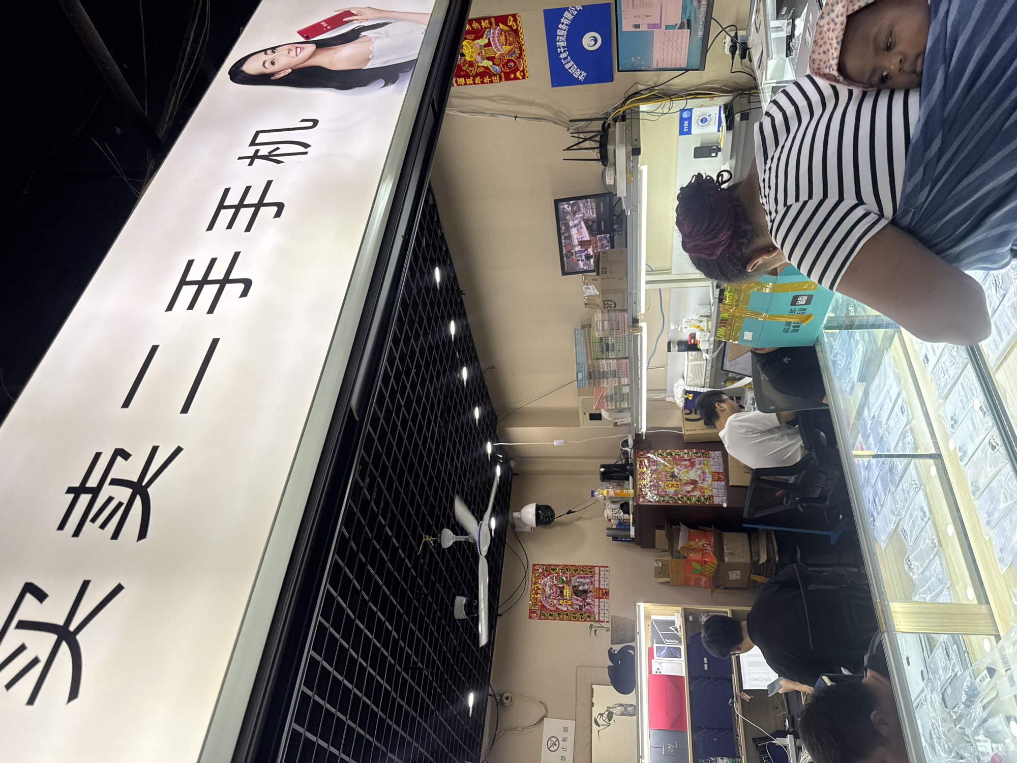 The image shows the interior of a mobile phone shop in Dadong District, China. A large illuminated sign above the shop reads "Buy and sell used mobile phones" in Chinese characters, with an image of a woman holding a phone. Inside the shop, several people are visible. Two men are working at desks, one of whom is looking at a computer screen. A woman with a baby wrapped in a sling is standing at a counter, looking at something on display. The shop is filled with shelves and display cases containing mobile phones and accessories. The environment appears to be a typical retail space, with fluorescent lighting and a black tiled ceiling.