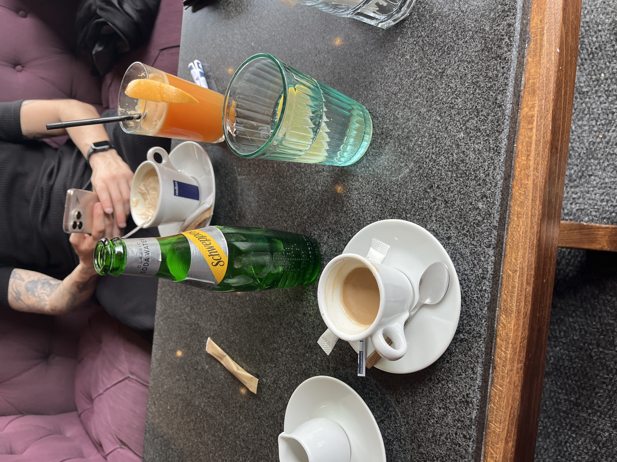 The image shows a table in a cafe in Varna, Bulgaria. A person is sitting on a purple cushioned bench, holding a smartphone. On the table, there is a glass of orange juice with a straw and orange slice, a turquoise glass with lemon slices, two empty coffee cups and one coffee cup with coffee, all on saucers, a green Schweppes soda water bottle, and a sugar packet.