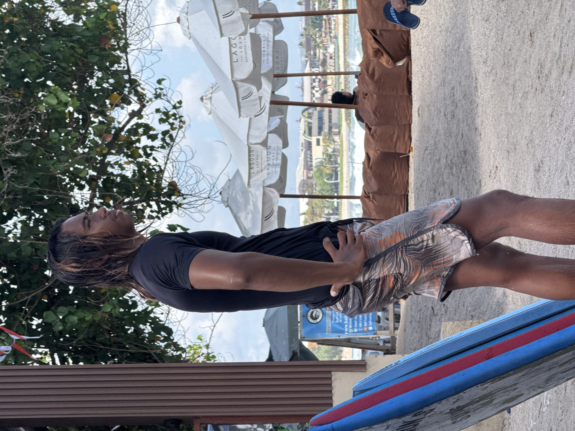 In Kuta, Indonesia, a young male with long, dark, wet hair stands on a sandy beach. He is wearing a black t-shirt and patterned swim shorts. He stands with his hands on his hips, looking to his right, towards the ocean. Beside him, to his left, is a surfboard with blue, red, and grey stripes. Behind him, several white beach umbrellas are set up, with the "Lagoon Lounge" logo visible on them. Beyond the umbrellas, a body of water with waves is visible, and in the distance, buildings and palm trees line the shore. The overall scene is that of a beach resort area.