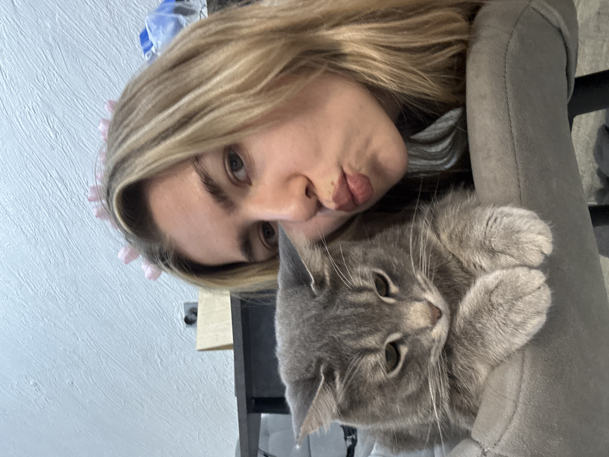 An indoor selfie captures a young woman and a gray tabby cat. The woman, with blonde hair and light-colored eyes, is seen from the chest up, her face angled towards the camera. She is making a "duck face" or pouting with her lips. A light pink, possibly floral, accessory is visible in her hair on the left side of her head. Her expression suggests playfulness or affection.
Next to her, the gray tabby cat rests, seemingly on a gray fabric chair or sofa armrest. The cat's head is visible, with its ears slightly perked and prominent whiskers. Its eyes are a striking green-amber color, looking towards the camera with a calm, relaxed expression. Its front paws are tucked together on the fabric.
The background features a light-colored, textured wall. Portions of dark furniture or objects are also faintly visible behind the woman and cat. The overall impression is one of a candid, affectionate moment between a pet owner and their cat. The image is taken at an angle, giving it a dynamic, casual feel. No text is visible in the image.