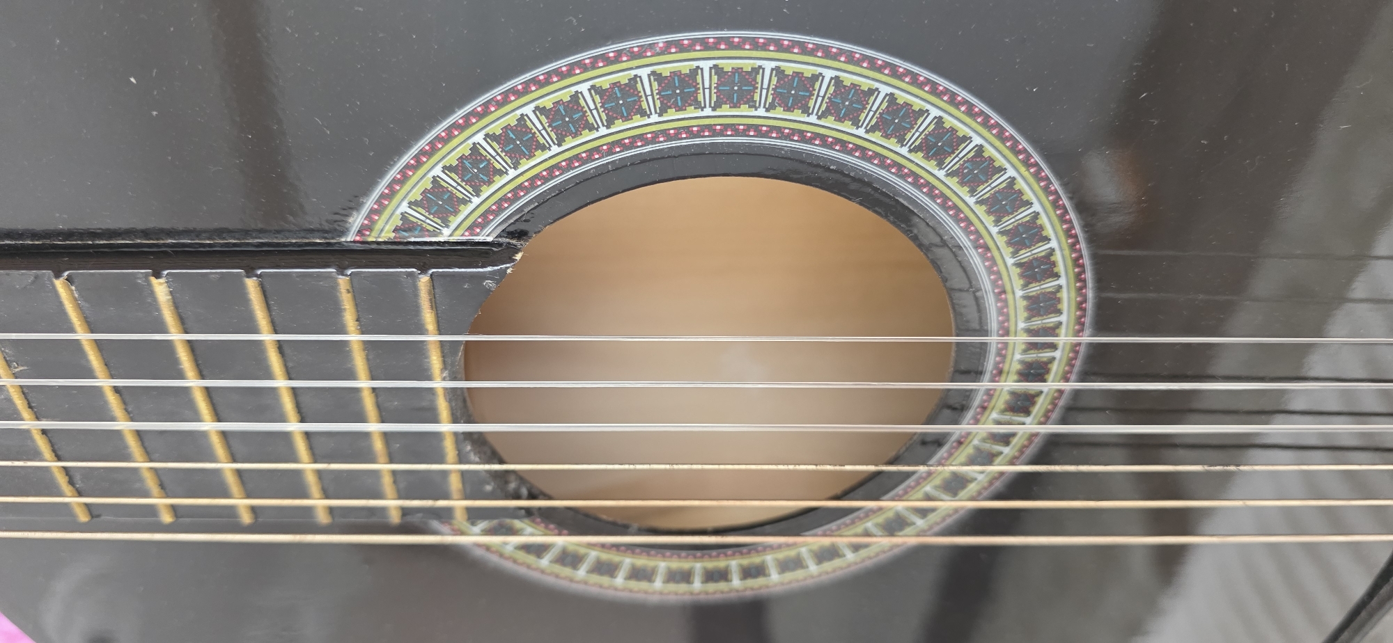 The image shows a close-up, eye-level, vertical view of the soundhole and strings of a black acoustic guitar. The soundhole is circular and decorated with an intricate rosette pattern featuring concentric rings of yellow, red, and a repeating geometric design in teal and red. Six strings, appearing to be nylon, run vertically across the frame, stretched over a dark fretboard with yellow binding. The guitar's body is a glossy black. The background is out of focus, suggesting a domestic or studio environment, and there is no indication of the city of Sumgayit, Azerbaijan, or any specific location within it.