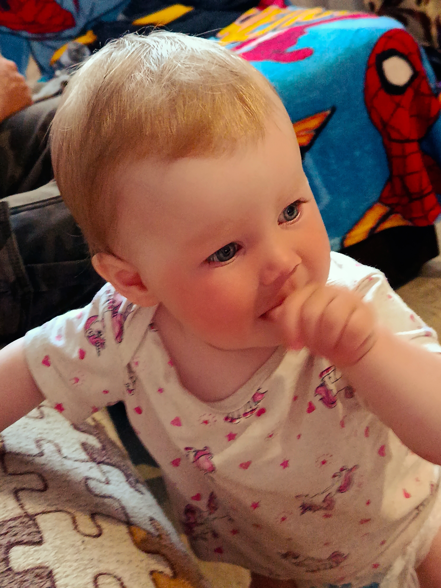 A baby with fair skin, light blue eyes, and reddish-blonde hair is the focal point of the image. The baby is wearing a white onesie patterned with pink unicorn motifs and hearts. The baby is holding their hand to their mouth, possibly sucking on their fingers.
In the background, a portion of a person wearing dark-colored pants is visible to the left. Behind the baby, there is a blue blanket with a Spider-Man design. The foreground shows part of a patterned blanket or rug.
The scene is indoors. No information on location in the city of Фоки, Russia, can be determined from the provided image.