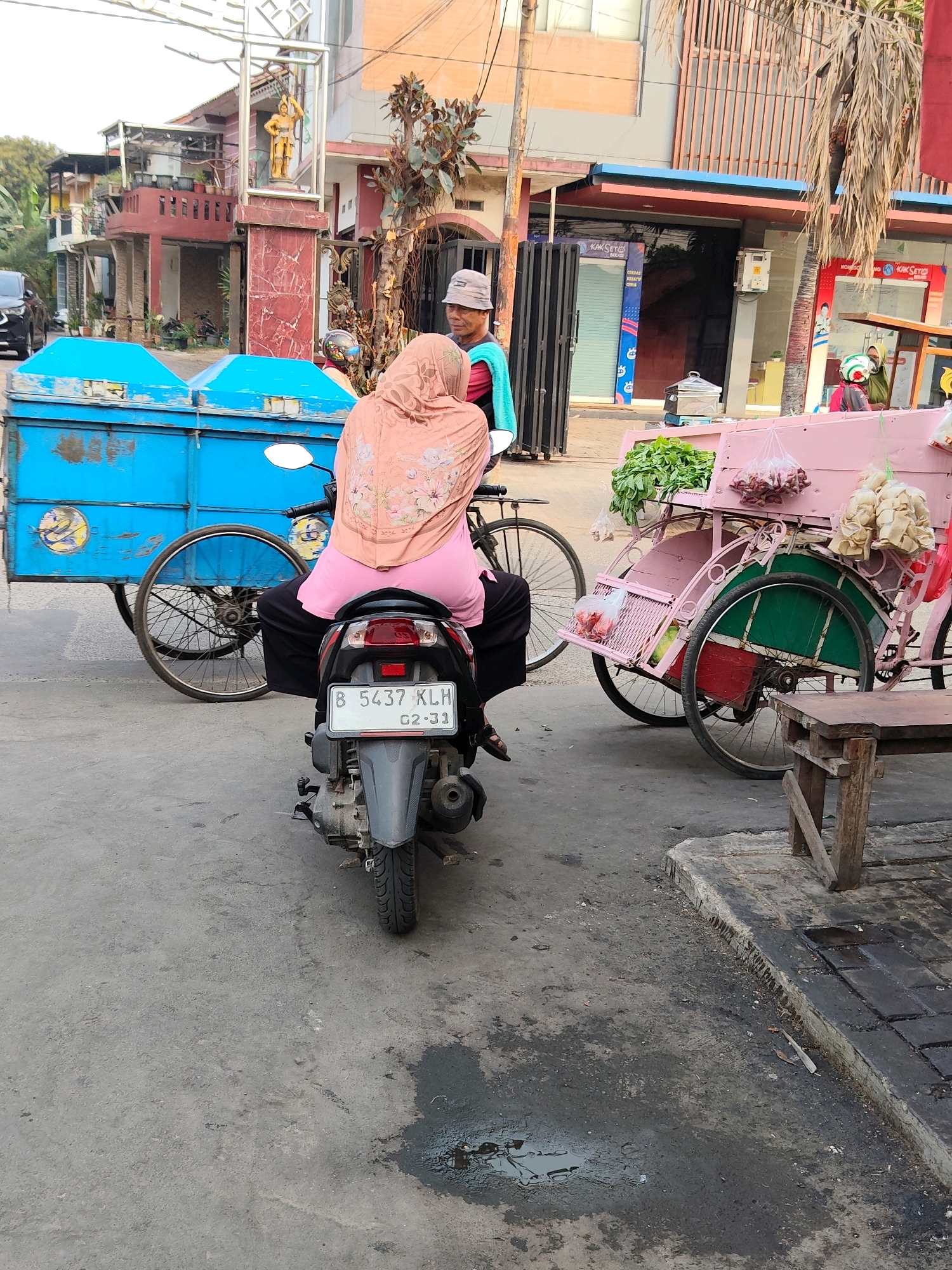 The image shows a street scene in Bekasi, Indonesia. In the foreground, a person wearing a pink headscarf and a pink shirt is seated on a black motorcycle, facing away from the camera. The motorcycle has a license plate that reads "B 5437 KLH 02-31". To the left of the motorcycle is a blue cart with two wheels. To the right of the motorcycle is a pink and green pedicab loaded with vegetables and what appear to be bags of food items. In the background, there are buildings, palm trees, and a statue of a golden figure. The ground is paved with asphalt and there are some puddles of dark liquid, likely water or oil.