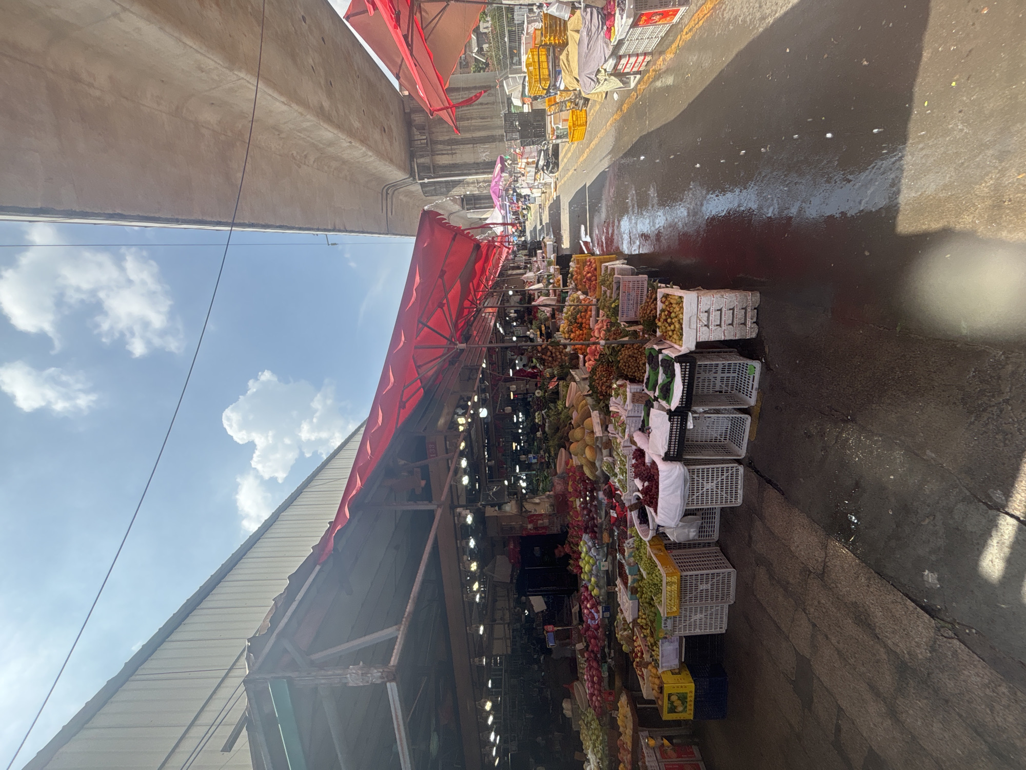 This image depicts an outdoor fruit market under an elevated highway structure in Nanhai District, China. The market stalls, covered by red awnings, are laden with a variety of fruits displayed in crates and baskets. The ground is wet and reflects the overhead structures and sky, suggesting recent rain or cleaning. In the background, more market stalls and various objects, including what appears to be a scooter, are visible. The sky is partly cloudy with bright sunlight.