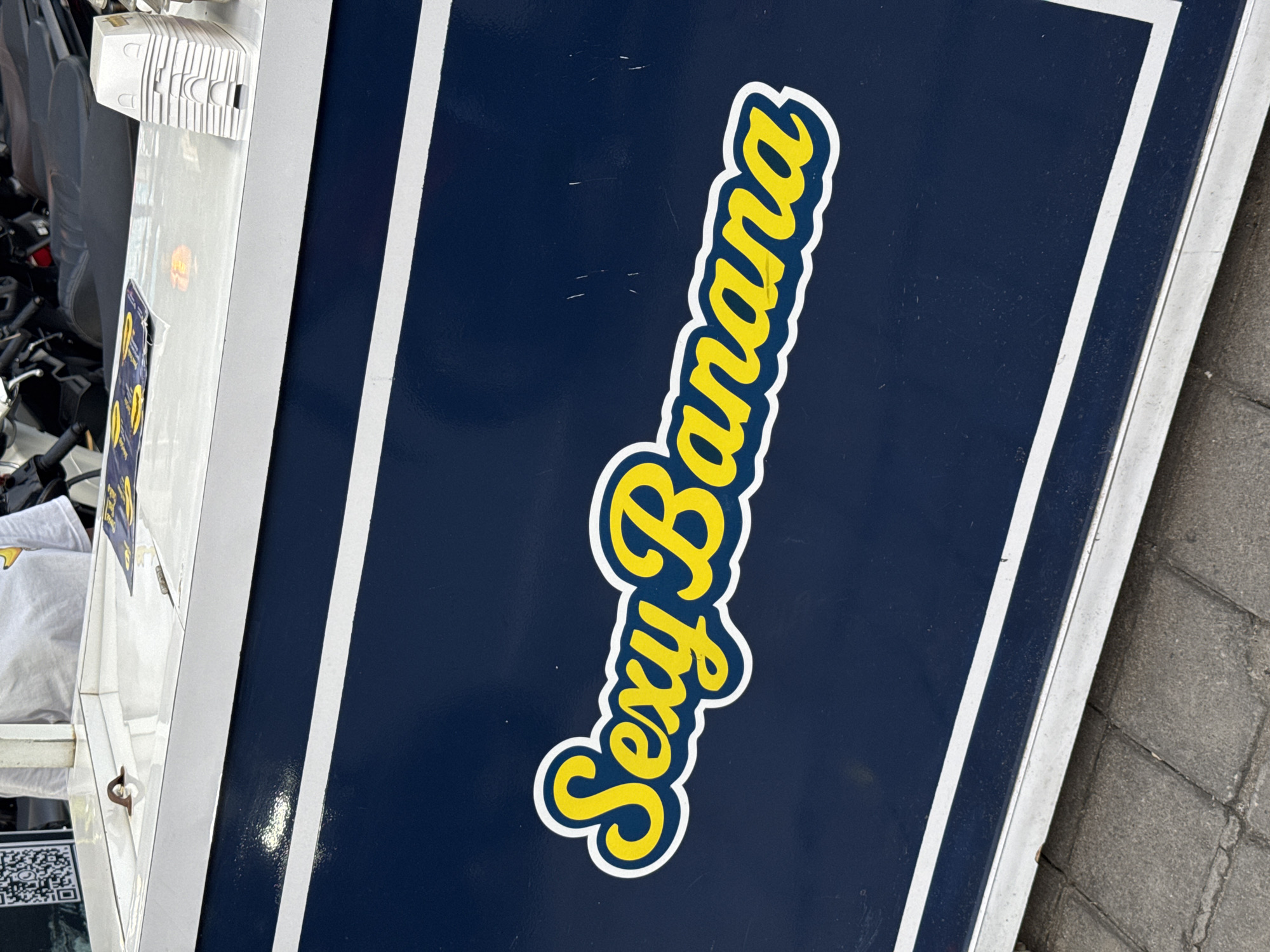 The image shows a portion of a vehicle, possibly a food truck, in North Kuta, Indonesia, as indicated by the QR code located near the bottom corner of the truck. The truck is dark blue with white trim. On the side, a sticker in yellow lettering with a white outline reads "Sexy Banana". Other objects are visible, including an object on the truck with many small, horizontal cuts. The image is taken at an angle, possibly at eye level. The background includes a sidewalk.