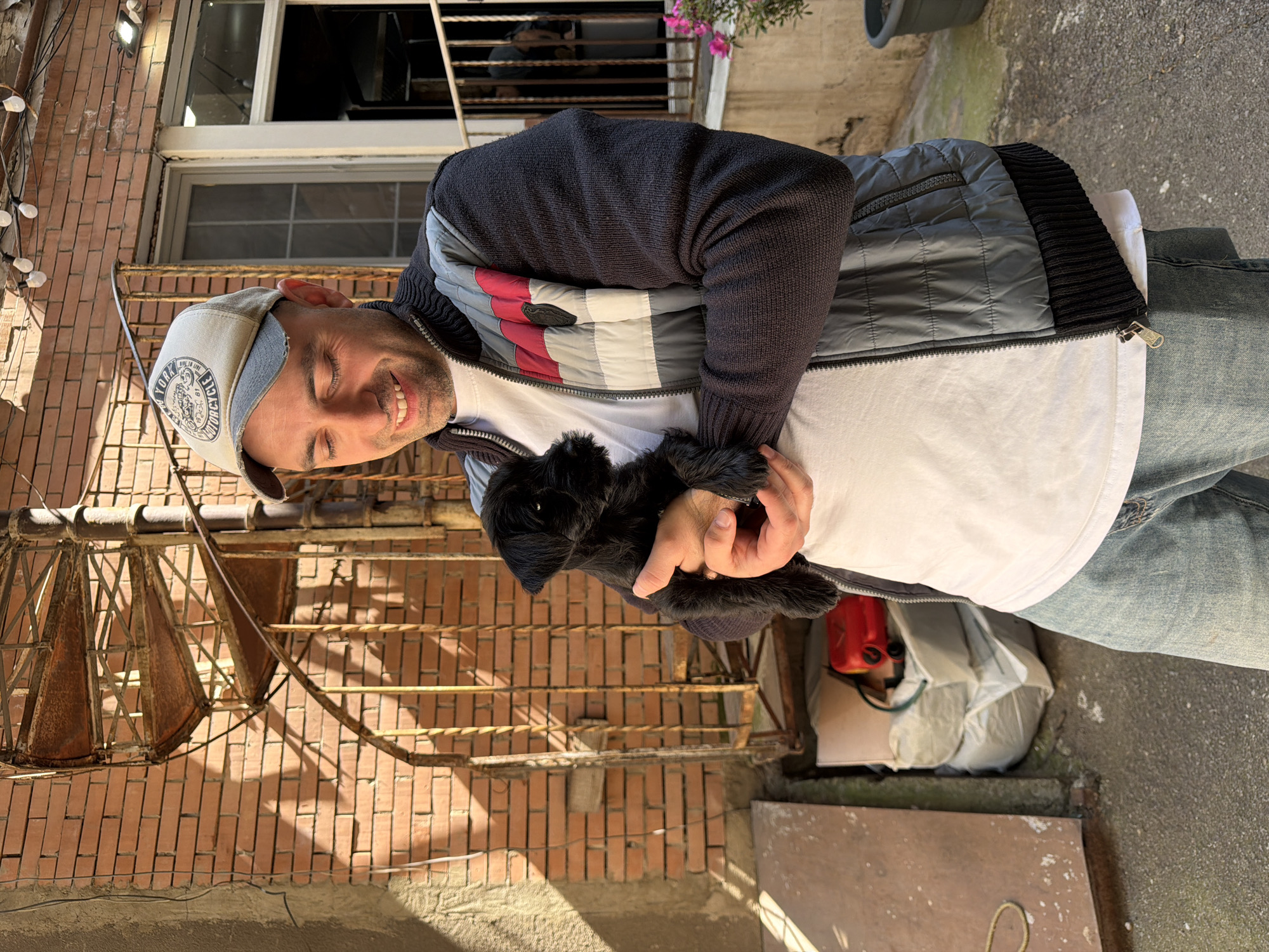 A man is captured in a sunny outdoor setting, holding a small black dog in his arms. He is smiling warmly, with his eyes closed or squinted, possibly due to bright sunlight. He wears a light-colored baseball cap with a "NEW YORK MOTORCYCLE" patch, a dark knitted jacket over a grey, red, and white striped quilted vest, a white t-shirt, and blue jeans. The small dog, which appears to be a black, fluffy terrier breed, is held gently against his chest.
The background features a reddish-brown brick wall, characteristic of older urban architecture. An ornate, rusty metal spiral staircase ascends along the wall on the left side of the frame, casting distinct shadows onto the bricks. Windows are visible on the building behind the man. String lights are draped along the wall to the left. In the upper right background, a potted plant with pink flowers adds a touch of color. The ground beneath the man's feet appears to be concrete or pavement, with some discarded white bags and a red container visible in the lower right corner. The strong sunlight and clear shadows suggest it is daytime, likely mid-morning or afternoon, and the weather is pleasant. The overall impression is one of a casual, affectionate moment between a pet owner and their dog in an urban backyard or alleyway in Tbilisi, Georgia.