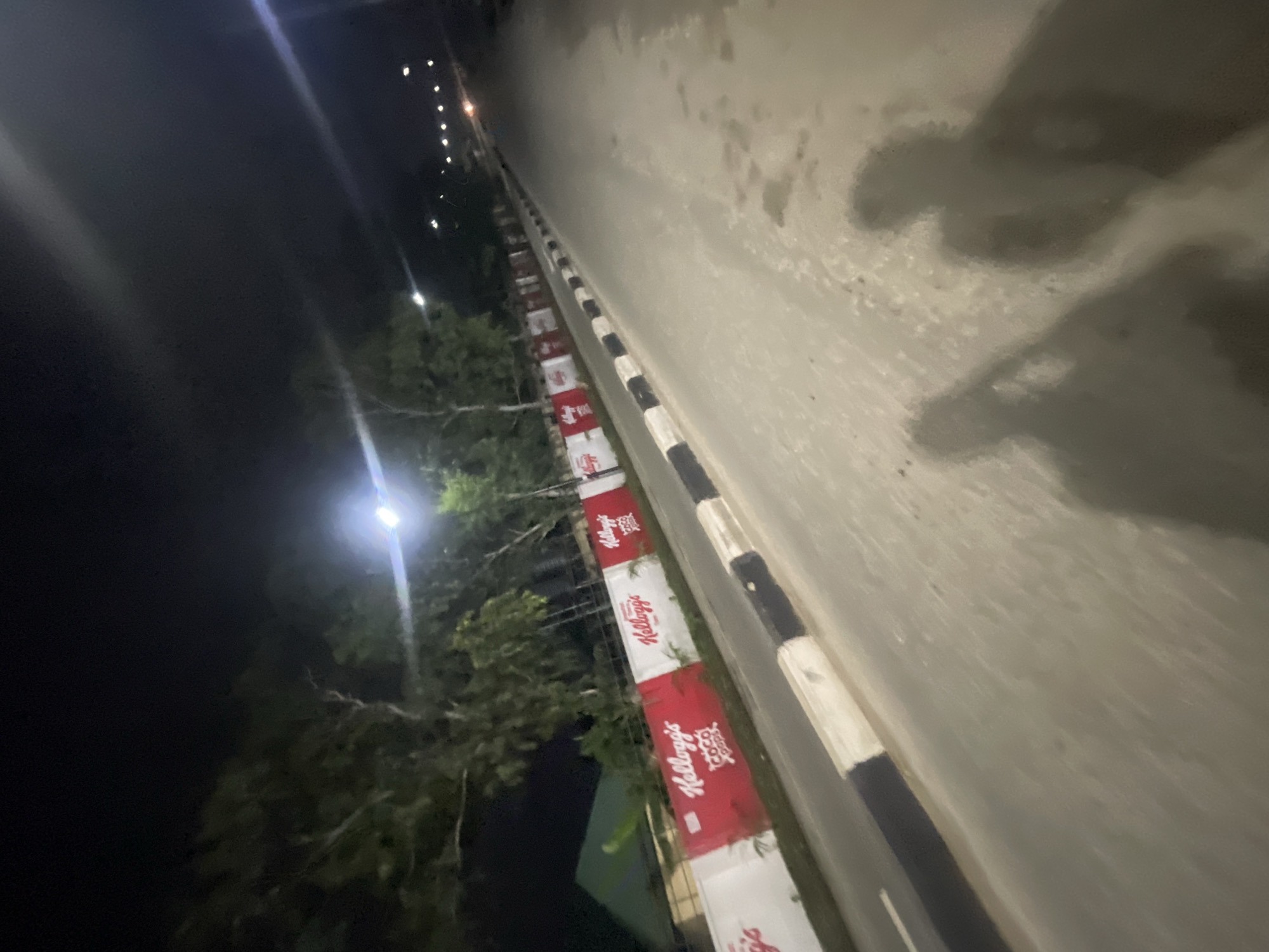 The image depicts a street scene at night in Akowonjo, Nigeria. A road with a black and white striped curb runs diagonally from the bottom left to the upper right of the frame. The road surface is wet and reflects the light from street lamps. On the left side of the road, there is a wall adorned with red and white banners that appear to be advertisements for Kellogg's Coco Pops. Behind the wall, trees and some buildings are visible. Two human shadows are cast on the road in the foreground, suggesting the presence of at least two people. The overall ambiance is that of a well-lit urban environment at night.