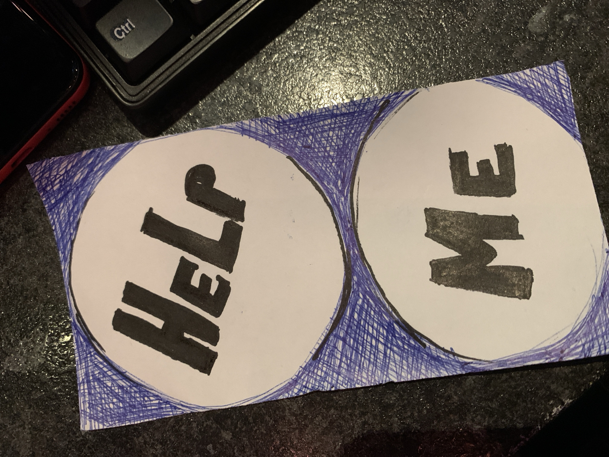 The image shows a handmade sign with the words "HELP ME" written in large black letters on two separate white circles. The circles are outlined in black and filled with blue crosshatching. The sign appears to be made of paper and is placed on a dark, textured surface, possibly a table. To the right of the sign, a portion of a black keyboard is visible, with a "Ctrl" key clearly legible. A red smartphone is also partially visible at the top right, resting on the surface. The lighting suggests an indoor environment, likely at night or in a dimly lit room. There is no discernible information about the location in Burgas, Bulgaria, in the image.
