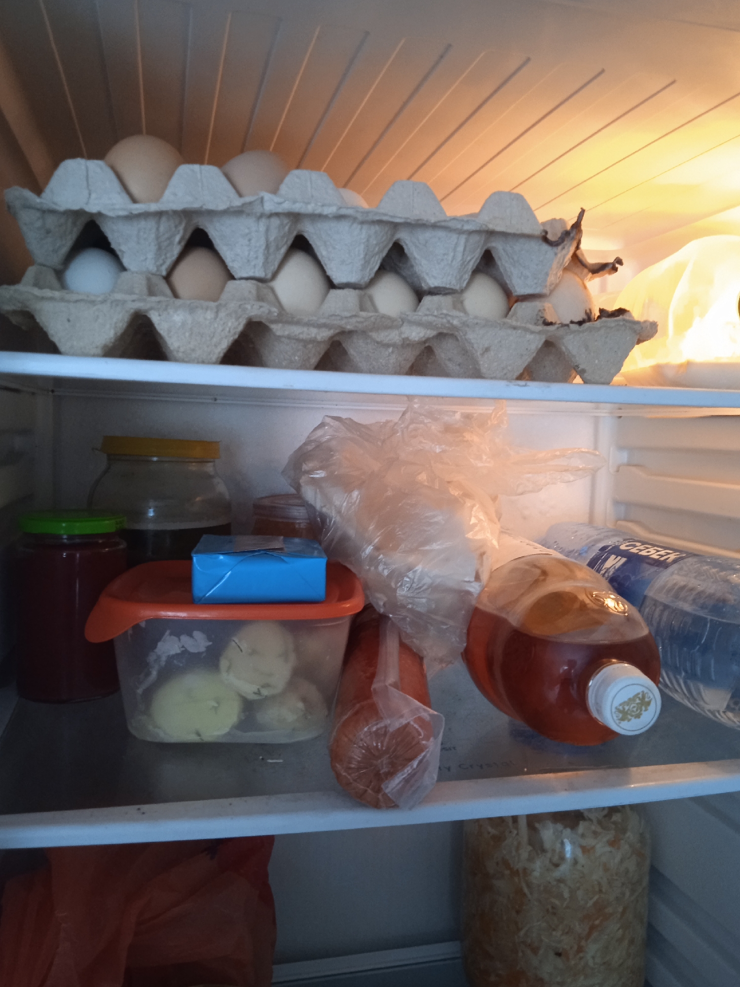 The image shows the inside of a refrigerator. On the top shelf, there are two cartons of eggs, with some eggs visible in the cartons. The shelf below contains several jars, some with green and red lids, and a transparent plastic container with what appears to be peeled potatoes. There is also a blue-wrapped package on top of the plastic container. Further to the right on this shelf, there is a plastic bag containing food items, a bottle of amber-colored liquid, and two bottles of water. The bottom shelf has a clear jar filled with a shredded, light-colored food item, and an orange plastic bag. The lighting suggests the refrigerator is illuminated from above and to the right. The scene is consistent with a domestic refrigerator located in Voznesensk, Ukraine.
