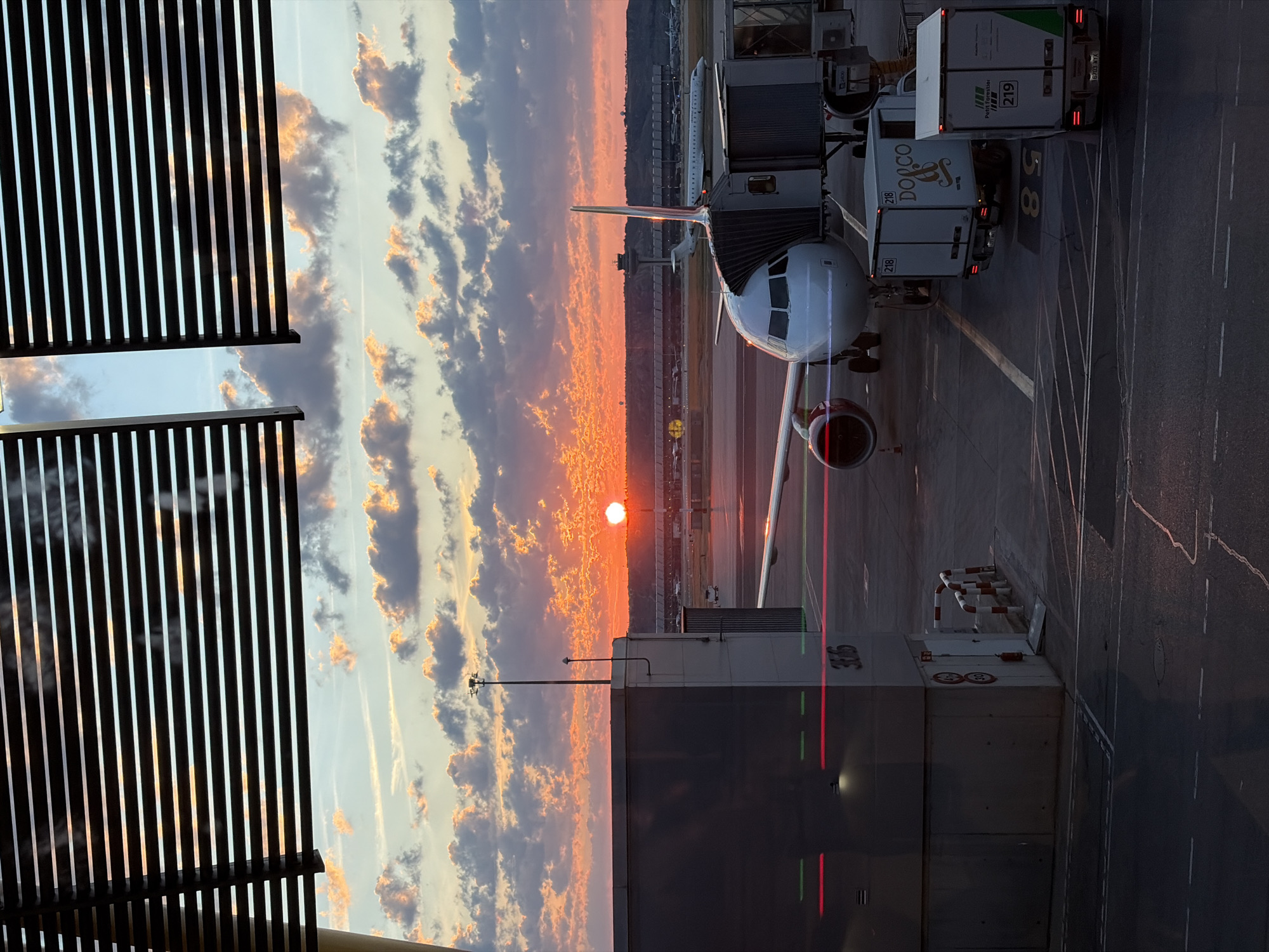 The image shows an airplane docked at a gate in an airport, with a sunset in the background. The airport is likely located near Madrid, Spain, given the landscape and architectural styles visible. The sun is setting on the horizon, with orange and purple hues dominating the sky. Clouds are scattered across the sky, catching the light of the setting sun.
An airplane is parked at the gate, with a catering truck attached. The plane is white with red accents. Airport infrastructure is present, including walkways, buildings, and signs. The boarding gate is connected to the plane, with people likely boarding or deboarding.
The overall environment is that of a busy airport during sunset, indicating a moment of transition as flights prepare for departure or arrival. The setting is likely Madrid, Spain.