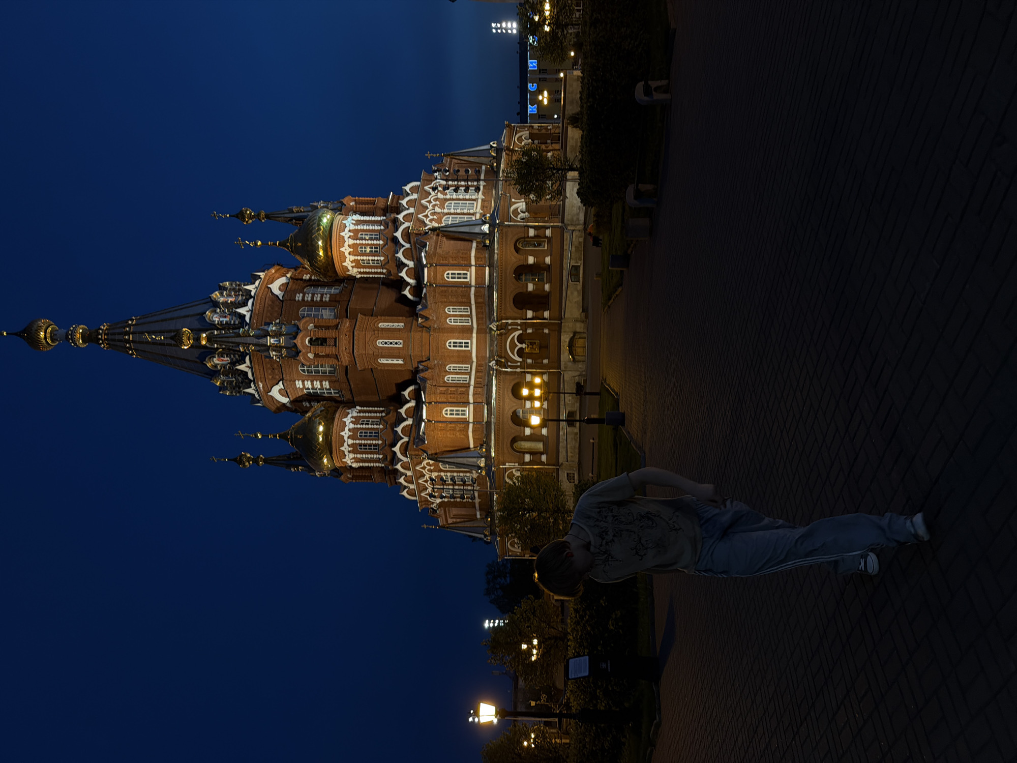 At night in Izhevsk, Russia, a person in a t-shirt and sweatpants is standing on a brick walkway in front of St. Michael's Cathedral. The cathedral is a brick building with white trim and multiple domes, illuminated by artificial light. The sky is dark blue, and streetlights are visible near trees along the walkway.
