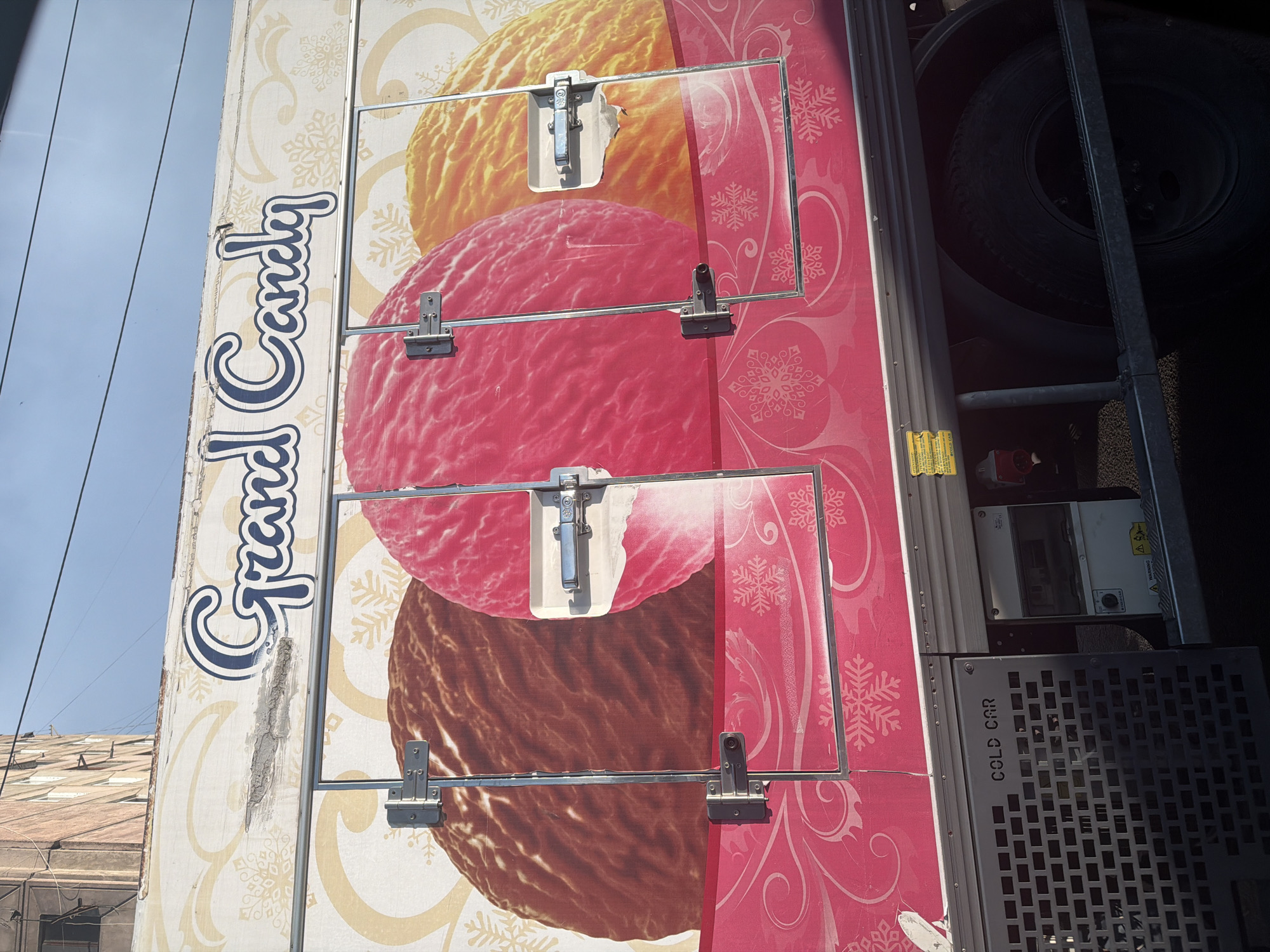 The image shows the side of a refrigerated truck with "Grand Candy" written in large blue and white letters across the top. The truck's side is decorated with large, stylized images of three scoops of ice cream in brown, pink, and orange. The lower portion of the truck features a pink graphic with white snowflake and swirl designs. There are metal doors with latches on the side of the truck, covering the ice cream images. Below the doors, a black metal grate with "COLD CAR" stenciled on it is visible, along with some electrical equipment. The truck is parked on a street, with a building and power lines visible in the background under a blue sky. The location is identified as Yerevan, Armenia.