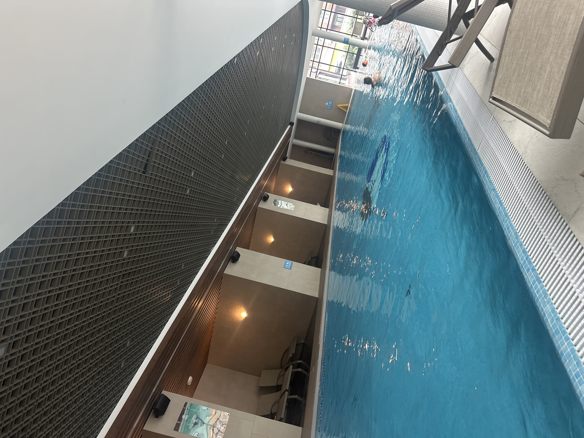 The image depicts an indoor swimming pool located in Estosadok, Russia. The pool is rectangular with blue water and a tile border. A person is swimming underwater, visible by their flippers. Another person is swimming on the surface of the water. Pool depth is indicated by signs along the side of the pool. Lounge chairs sit at the edge of the pool. One side of the pool is adjacent to a wall with a lattice design. There is also a white wall.
