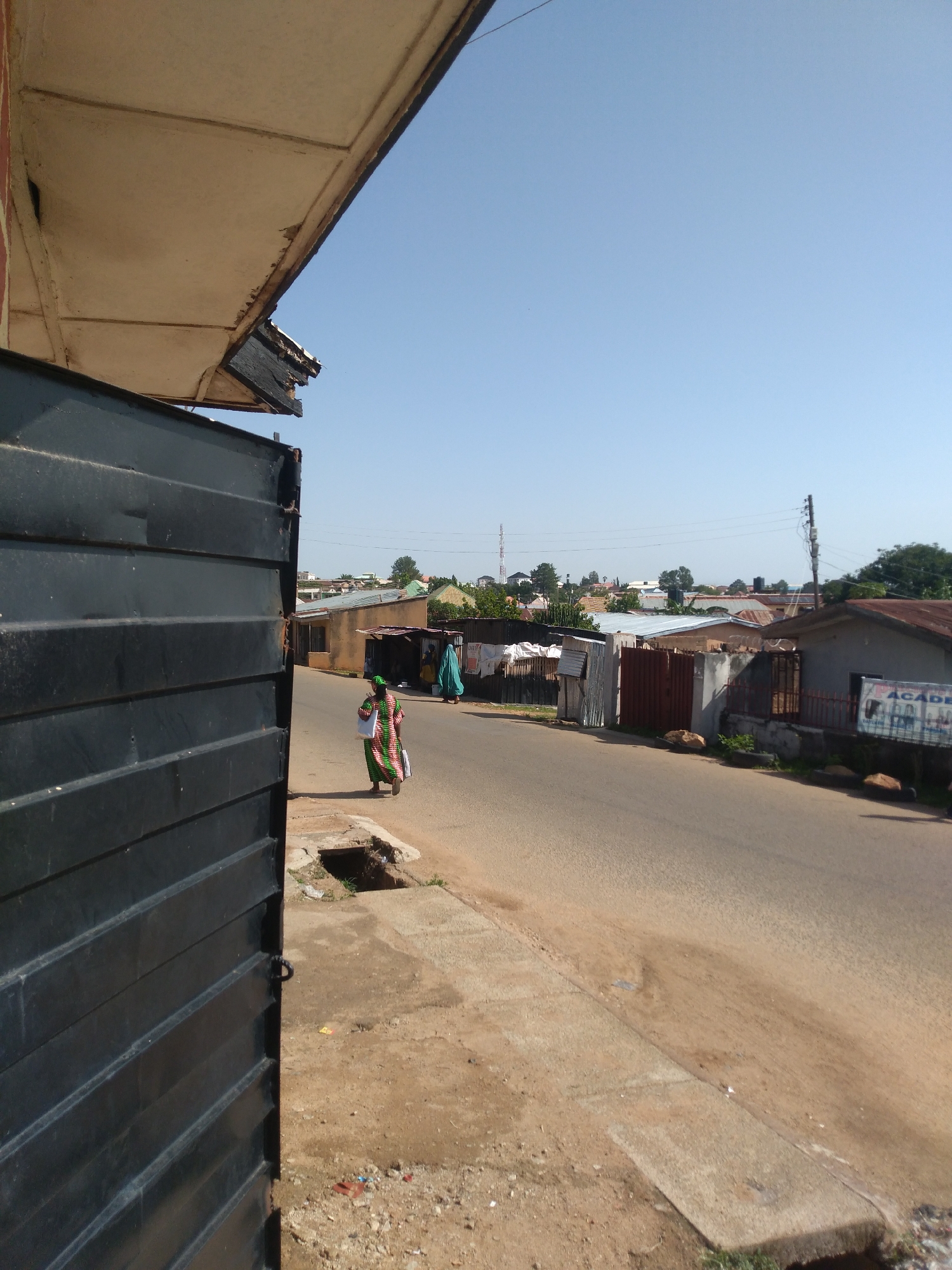 The image depicts a street scene in Jos, Nigeria, under a clear blue sky. In the foreground, a large, dark, corrugated metal gate dominates the left side of the frame, with a portion of a weathered overhang visible above it. A paved sidewalk runs alongside the gate, transitioning to a dirt and gravel path in places. A drainage grate is visible on the sidewalk.
The street itself is paved and appears to be a thoroughfare with modest buildings lining either side. On the right side of the street, a structure with a sign that reads "ACADEMY" is visible, along with other houses with corrugated iron roofs and various fencing. On the left side of the street, a woman in a green and pink patterned dress is walking away from the camera, carrying white bags. Another person in a blue garment is further down the street, near a group of buildings. The environment suggests a residential or mixed-use area within the city.