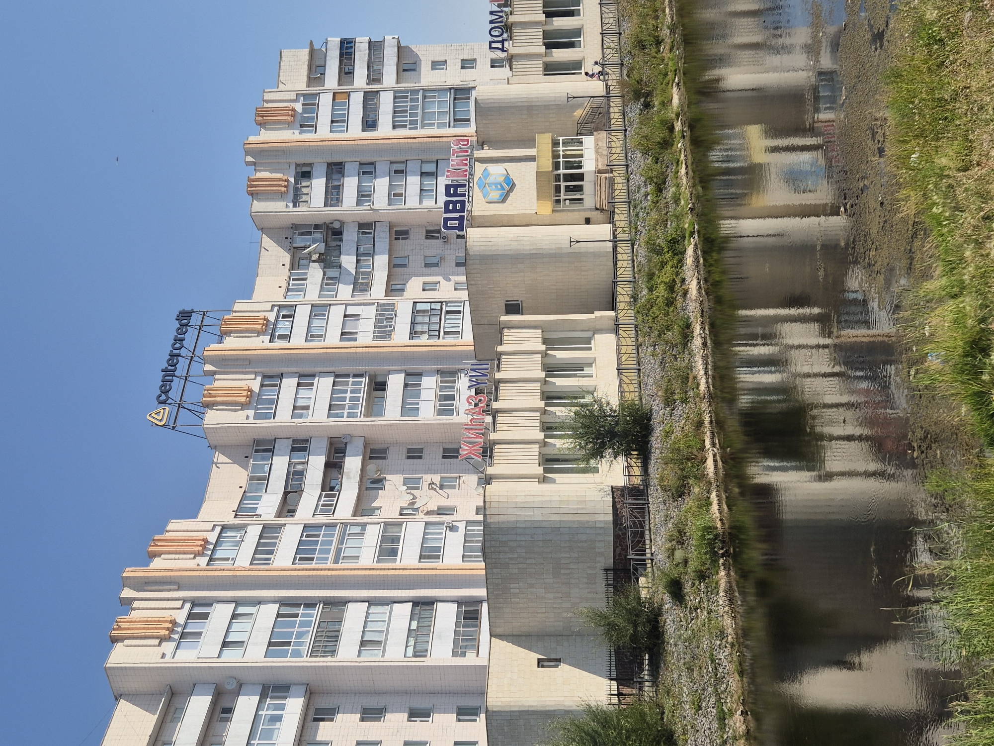 This is a photograph of a modern apartment building in Kokshetau, Kazakhstan, on a clear, sunny day with a blue sky. The building is multi-story with a light-colored facade, accented with decorative elements in orange and vertical cream-colored columns. Several signs are visible on the building. At the top, the sign "centercredit" is illuminated. On the lower part of the building, signs read "ЖИНаЗ УЙ" in red letters and "ДВА Кита" in blue and red letters. A logo featuring a blue and white cube with geometric shapes is also present.
In front of the building, there is a wide body of water, likely a river or canal, reflecting the building and the sky. The water's edge is lined with green grass and some rocky areas. A black metal fence runs along the bank between the building and the water. Two people on bicycles are visible near the entrance of the building. The overall scene depicts an urban environment with a focus on the architecture and its reflection in the water.
