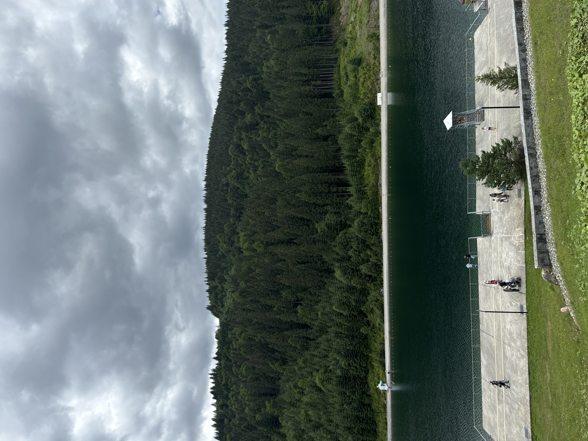 The image depicts a scenic view of a lake and a forested mountain in Polianytsia, Ukraine. The lake is a deep green color and appears calm, with small waves gently lapping at the shore. Along the edge of the lake, a concrete walkway or platform is visible, with several people walking and standing on it. There is also a lifeguard tower with stairs leading up to it. A dense forest of evergreen trees covers the mountainside that rises behind the lake. The sky is overcast with heavy, gray clouds, suggesting a possibly cool or rainy day. The foreground shows a grassy slope with some rocks.