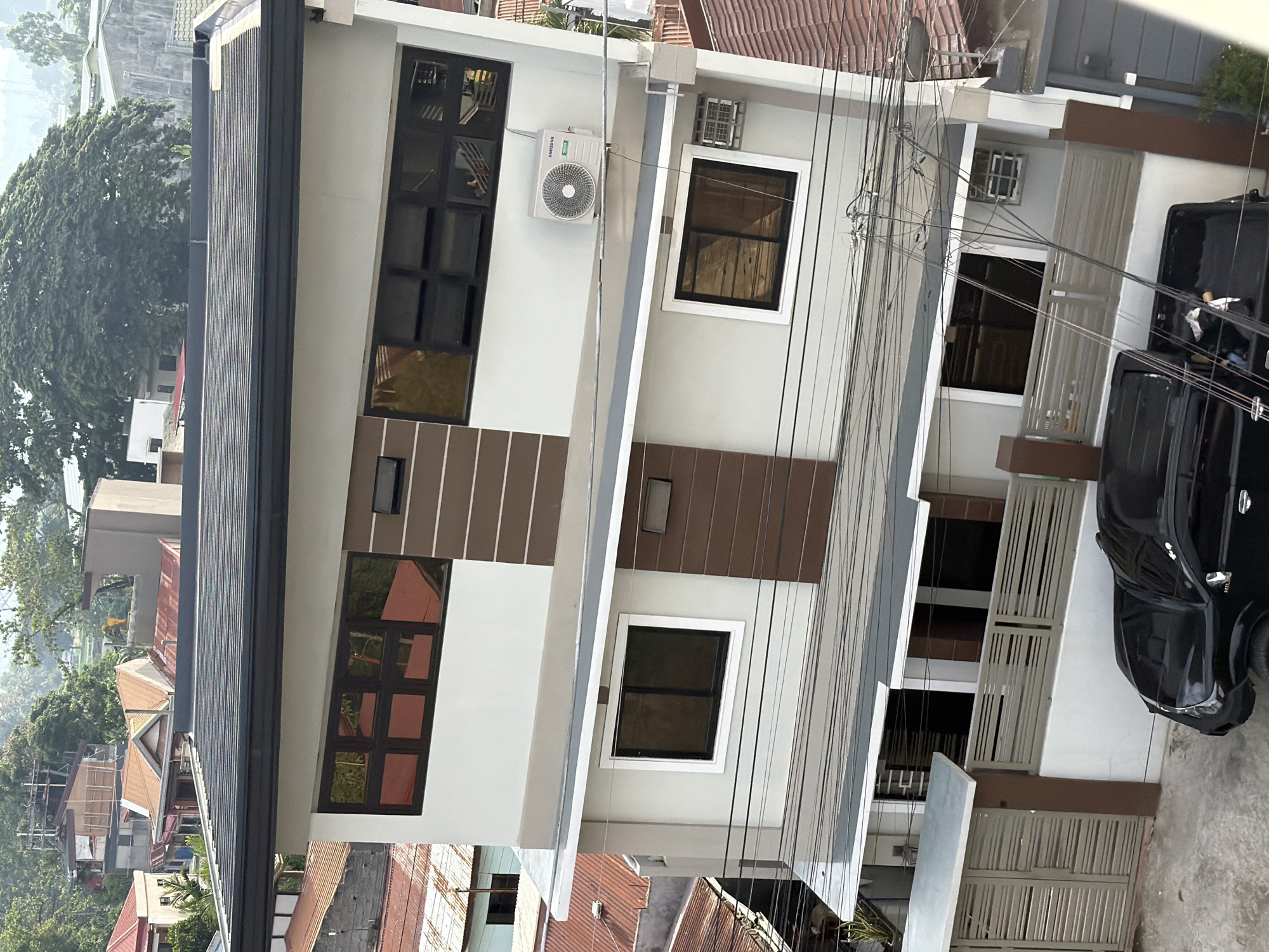 The image presents a residential building in Davao City, Philippines. The structure is a multi-story building with a white and brown facade, featuring rectangular windows with dark frames. An air conditioning unit is mounted on the exterior wall. Various electrical wires are visible, crisscrossing in front of the building. In the foreground, part of a black vehicle is visible. The surrounding environment includes other buildings and lush green trees. The overall setting suggests a densely populated urban area.