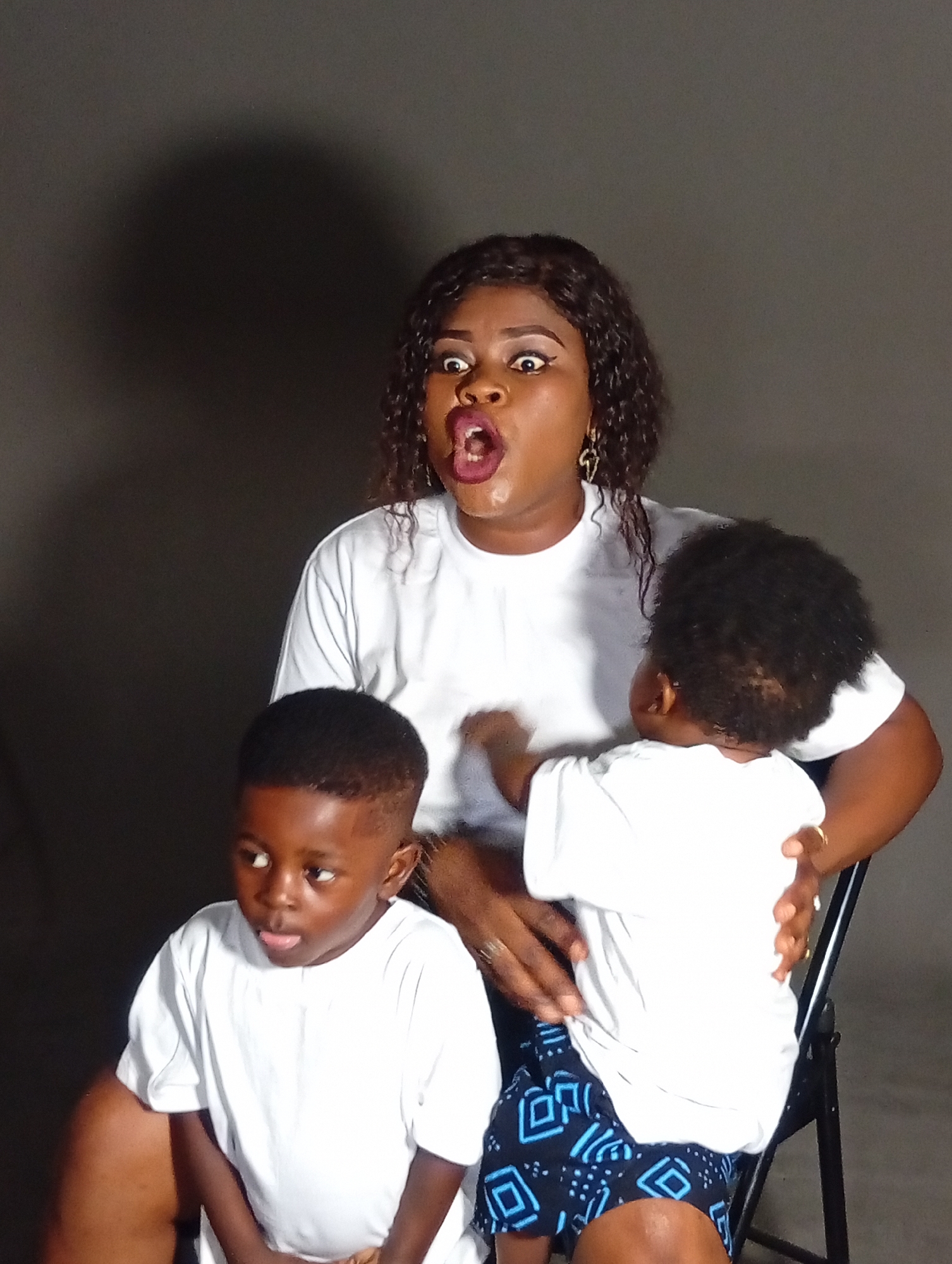 A woman with dark curly hair and a white t-shirt sits on a chair, her mouth open in surprise. She is holding two young children. A boy in a white t-shirt is in the foreground, looking to the left with his tongue sticking out. Another child, also in a white t-shirt, is being held by the woman, facing away from the camera. The background is a plain dark gray.