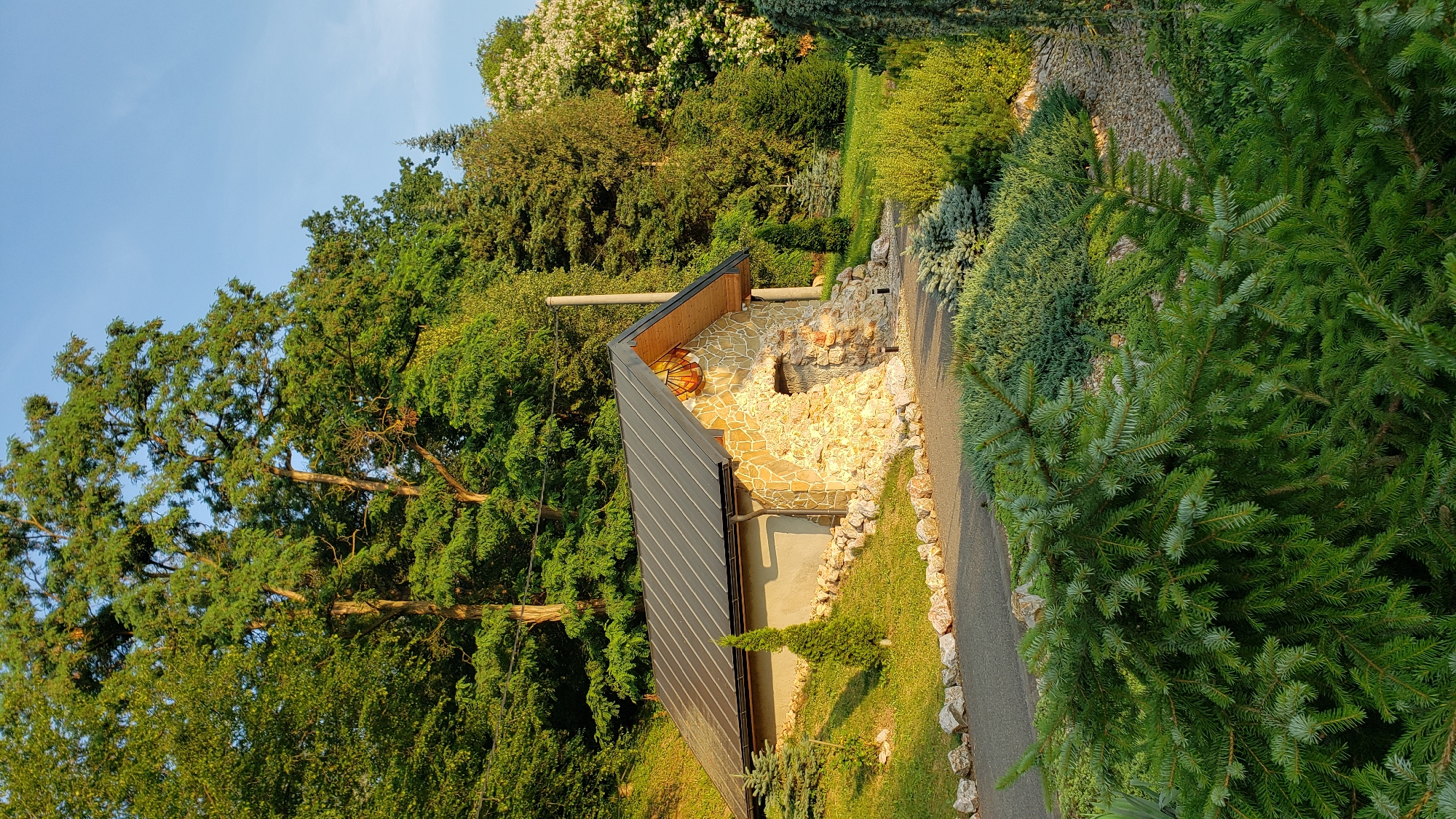 A small stone structure with a dark metal roof is nestled into a grassy hillside, surrounded by lush greenery. The building has a rough stone facade with an arched opening. A pathway made of grey asphalt and bordered by light-colored stones leads to the structure. Various coniferous trees and shrubs are visible around the building and along the path. The scene is bathed in warm sunlight, suggesting it is either morning or late afternoon. The sky is a clear, light blue. The image is taken from a slightly elevated perspective, looking towards the building and the surrounding landscape. The presence of a pole in the background might indicate the area is near some form of infrastructure, but it is not definitively identifiable within the context of Prešov, Slovakia, based solely on visual cues.