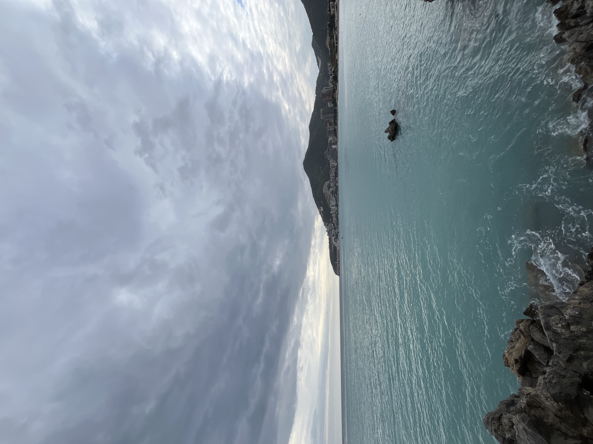 The image shows a seascape in Budva, Montenegro. The view is dominated by the turquoise water of the Adriatic Sea, which stretches from the foreground to the horizon. Dark, rocky cliffs are present in the lower right corner, with waves crashing against them. Further along the coastline, a mountain slopes down towards the sea, with buildings lining its base. The sky is overcast with dense clouds, ranging in color from light grey to dark grey. The horizon is visible as a line where the sea meets the sky. A few small rocks jut out of the water in the distance. No people are visible in the frame.