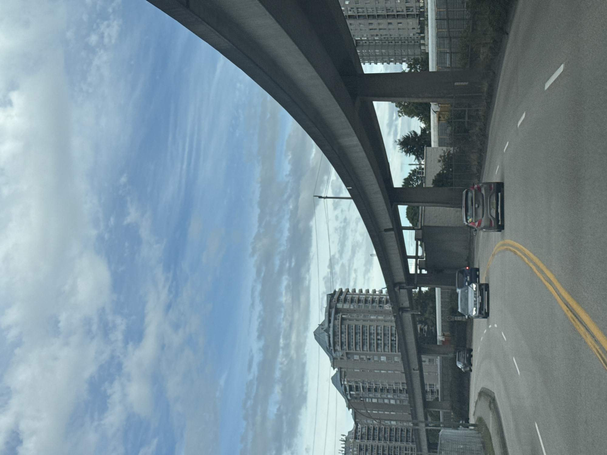 The image shows a road with several cars driving under an elevated highway structure. In the background, there are tall buildings. The sky is cloudy. The location is identified as New Westminster, Canada.
The road is paved and has lane markings, including double yellow lines and dashed white lines. A grey sedan is in the foreground, followed by a silver Volkswagen sedan and a dark blue SUV. Further back, a black SUV is visible.
The elevated highway is a concrete structure supported by pillars. Buildings are visible on either side of the highway. On the left, there are several tall, modern apartment buildings with distinctive peaked roofs. On the right, another tall building, likely an apartment or office building, is partially visible.
The environment appears to be urban, with infrastructure for transportation and residential or commercial buildings. The weather seems overcast with scattered clouds.