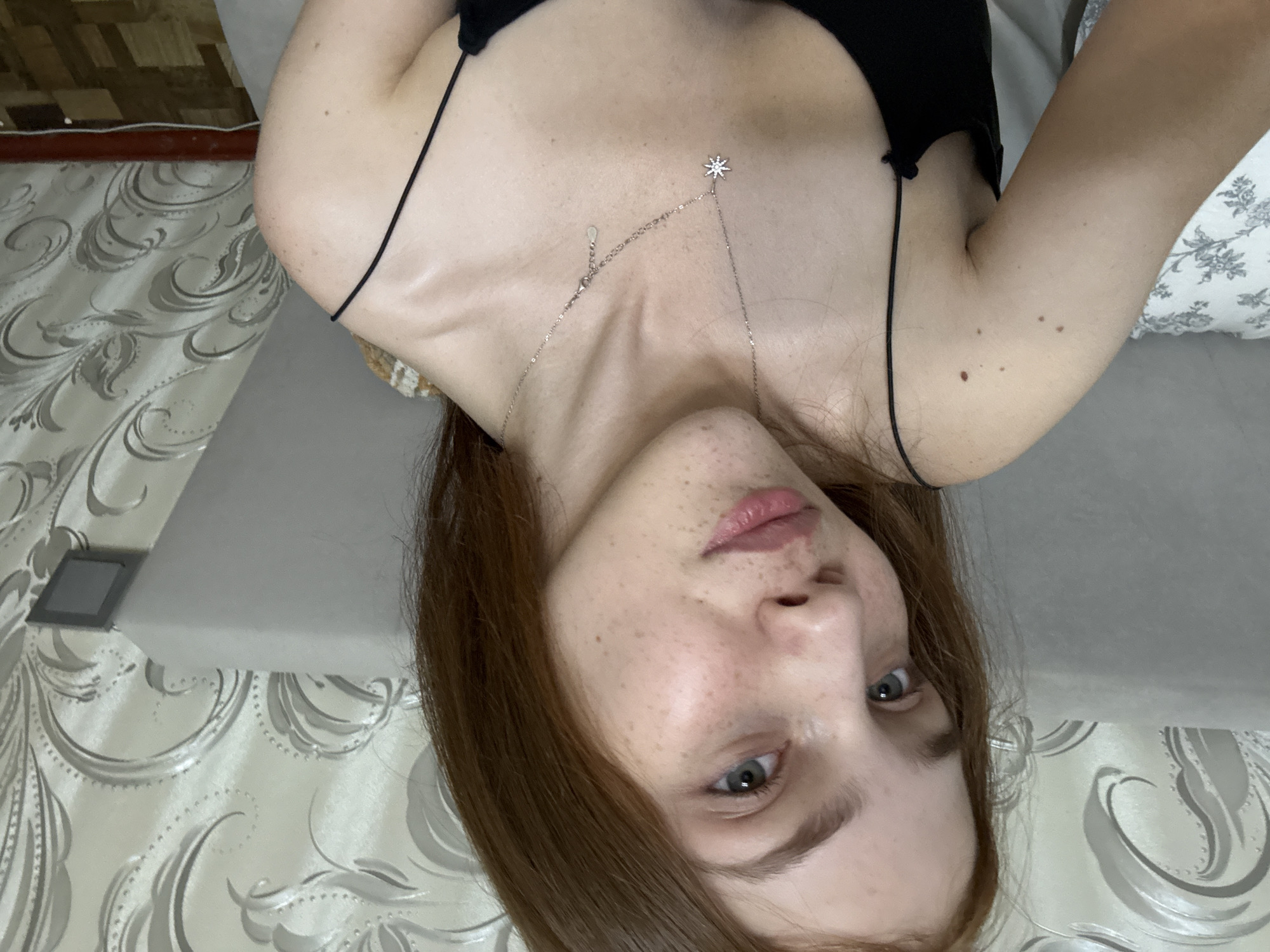The image is a close-up, selfie-style photograph of a young woman with fair skin, freckles, and long auburn hair. She is wearing a black spaghetti strap top and a silver necklace with a star-shaped pendant. The background consists of a textured wallpaper with a swirling pattern and a portion of a light-colored headboard. A pillow with a floral pattern is visible to the left. The lighting appears to be natural or from an indoor source, casting a soft glow on her face. The scene suggests an indoor setting, possibly a bedroom. There is no information within the image to ascertain a specific location in Naberezhnye Chelny, Russia.