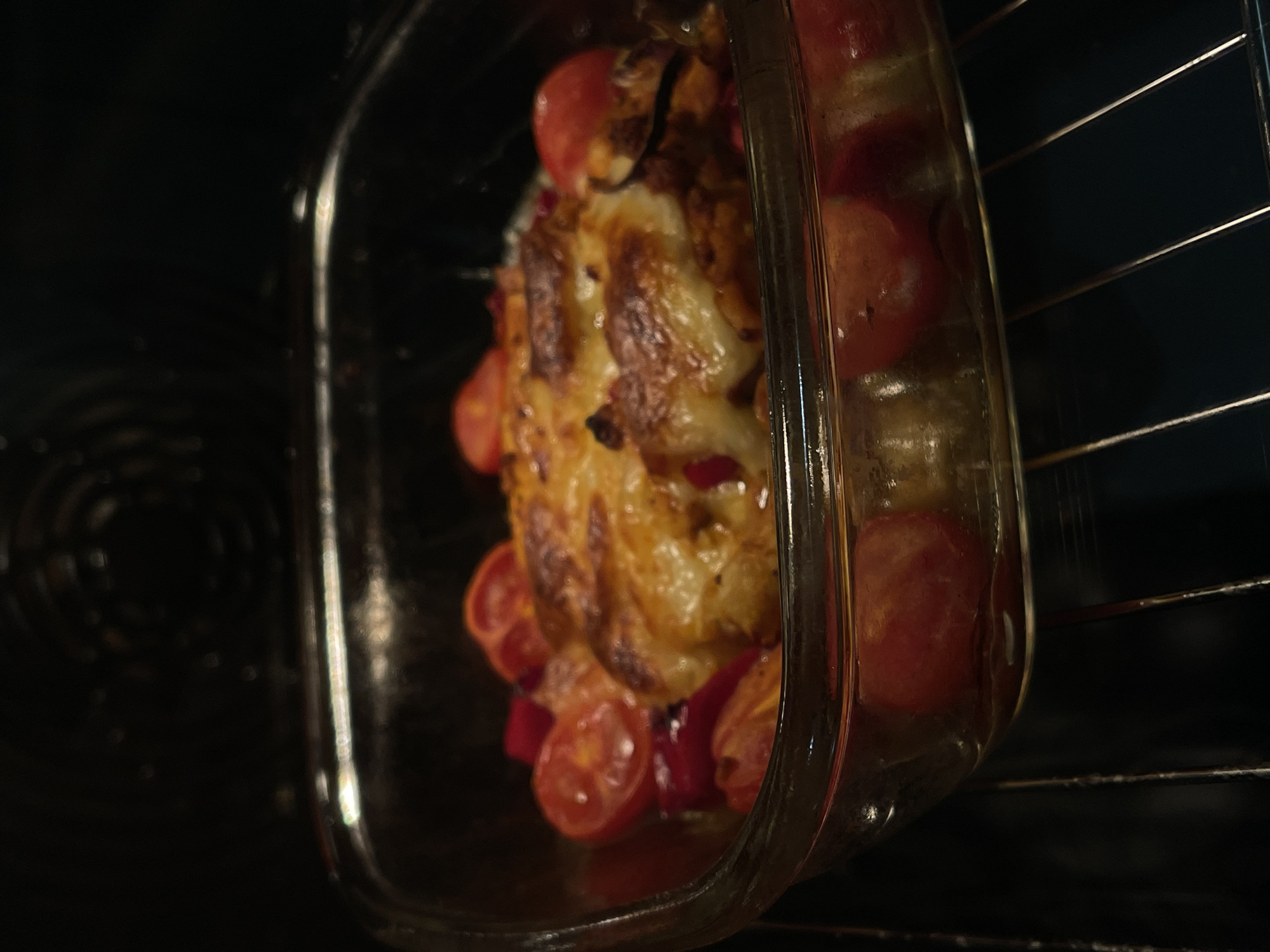 In a glass baking dish, a chicken breast is baked with halved cherry tomatoes and what appear to be red bell pepper slices. The chicken is golden brown and covered in melted, bubbly cheese. The dish is placed on an oven rack, inside a dark oven with the heating element visible in the background. The lighting is dim, suggesting the oven door is closed. The scene appears to be in a kitchen.