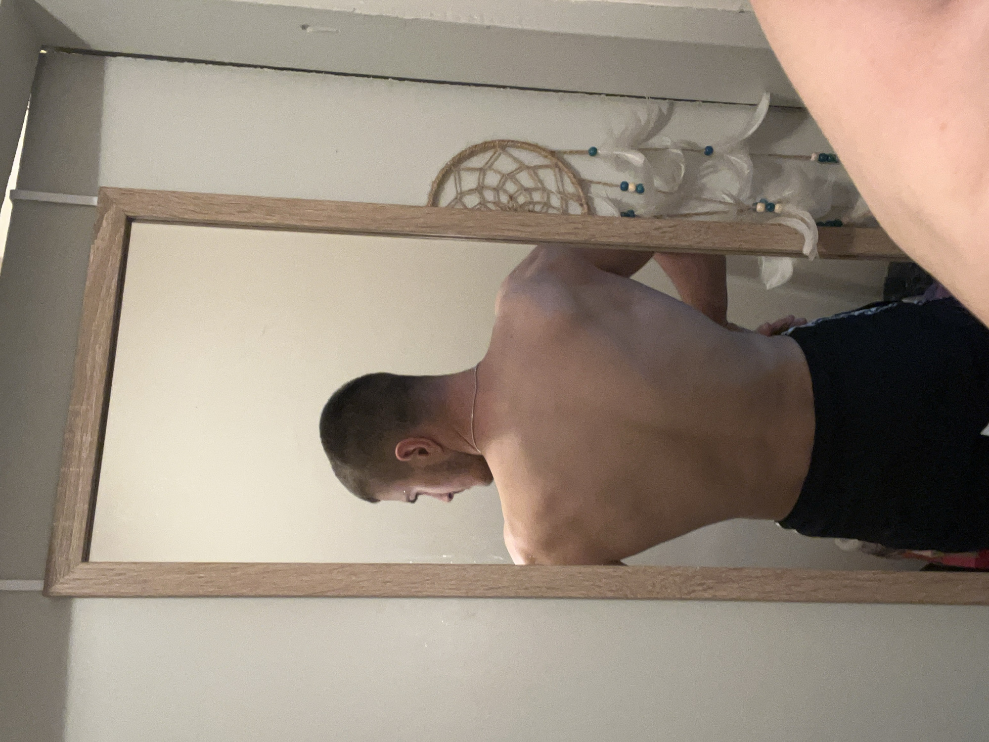 The image shows a person reflected in a rectangular mirror with a light brown wooden frame. The person has short brown hair and is wearing black shorts. The upper part of their body is bare. Behind the mirror, a dreamcatcher is attached to the wall. The room appears to be indoors, with white walls. The image's location is unidentified.