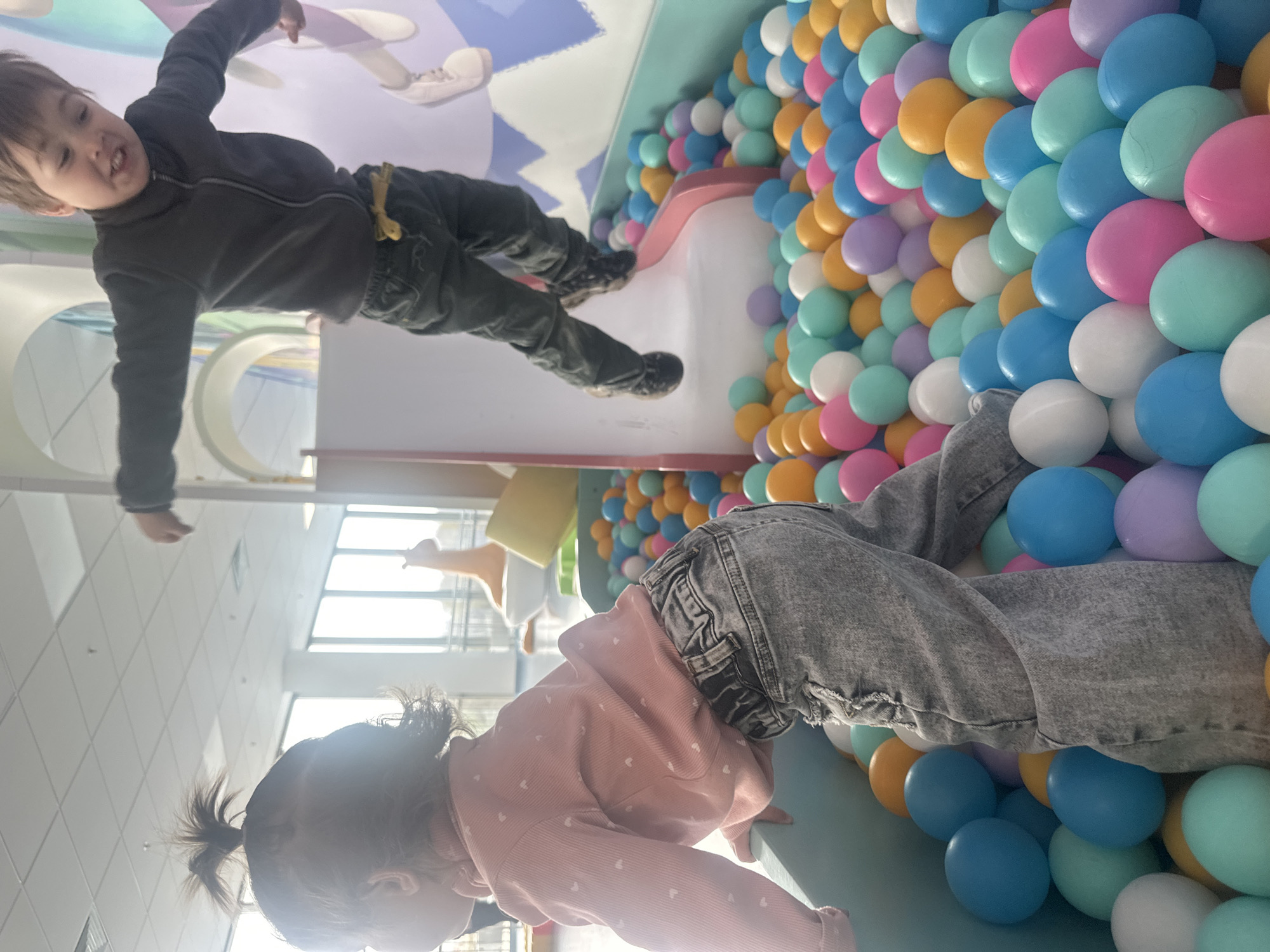 The image captures a vibrant indoor play area in Khimki, Russia, featuring two young children engaged in playful activities. The scene is brightly lit, suggesting daytime, with natural light streaming in from large windows visible in the background, hinting at a clear day outside.
In the upper left, a young boy is prominently featured, appearing to walk upside down due to a reflective surface beneath him. He wears a dark grey zip-up jacket, dark pants with a yellow accessory clipped to the side, and dark shoes. He looks towards the camera with an excited, open-mouthed expression, revealing his teeth. The reflective surface creates an illusion where he seems to defy gravity.
To the right, a large section of the frame is filled with a wall or play structure densely packed with numerous pastel-colored plastic balls. These balls come in various shades, including pink, light blue, purple, yellow, and white, creating a cheerful and inviting atmosphere. A young girl, seen from behind, is actively engaging with this ball pit. She has dark hair tied in a small ponytail and is dressed in a light pink long-sleeved top adorned with small white heart patterns, paired with grey ripped jeans. She is leaning or crawling forward, with her lower body partially submerged among the colorful balls.
The setting is clearly a children's soft play or entertainment center, characterized by its bright colors and child-friendly design elements. There is no visible text in the image.