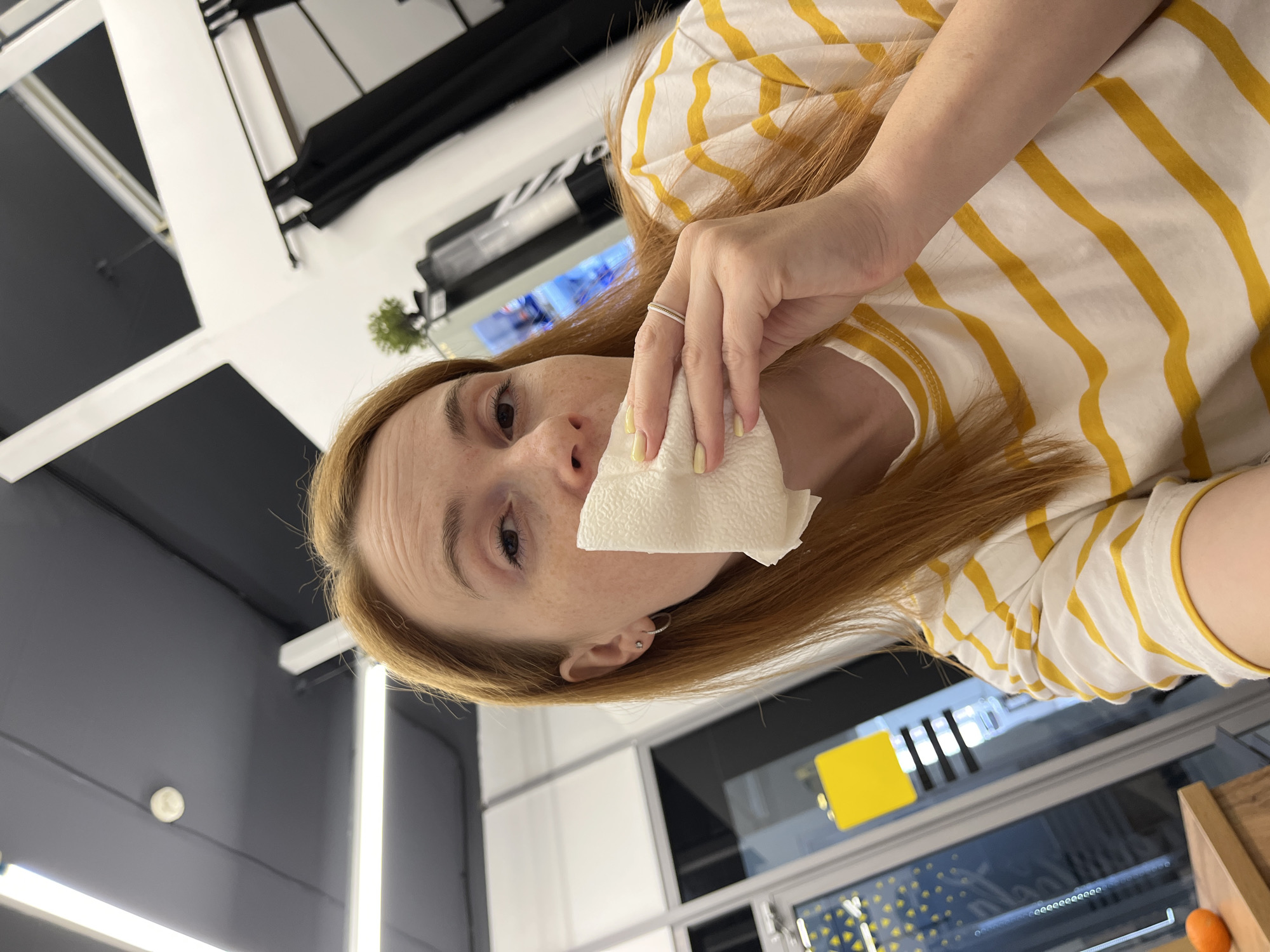 A woman with reddish-blonde hair, wearing a striped yellow and white shirt, is holding a tissue to her nose. She has pale skin and is wearing a ring on her left ring finger. She appears to be inside a building with a modern interior, featuring grey and white walls, and a complex ceiling structure with black and white elements. In the background, there is a glimpse of a window or glass partition with a visible design. Given the request for the location, it's a presumption based on the query that this scene is taking place in Chaikovsky, Russia. However, this detail can't be visually confirmed from the image alone.