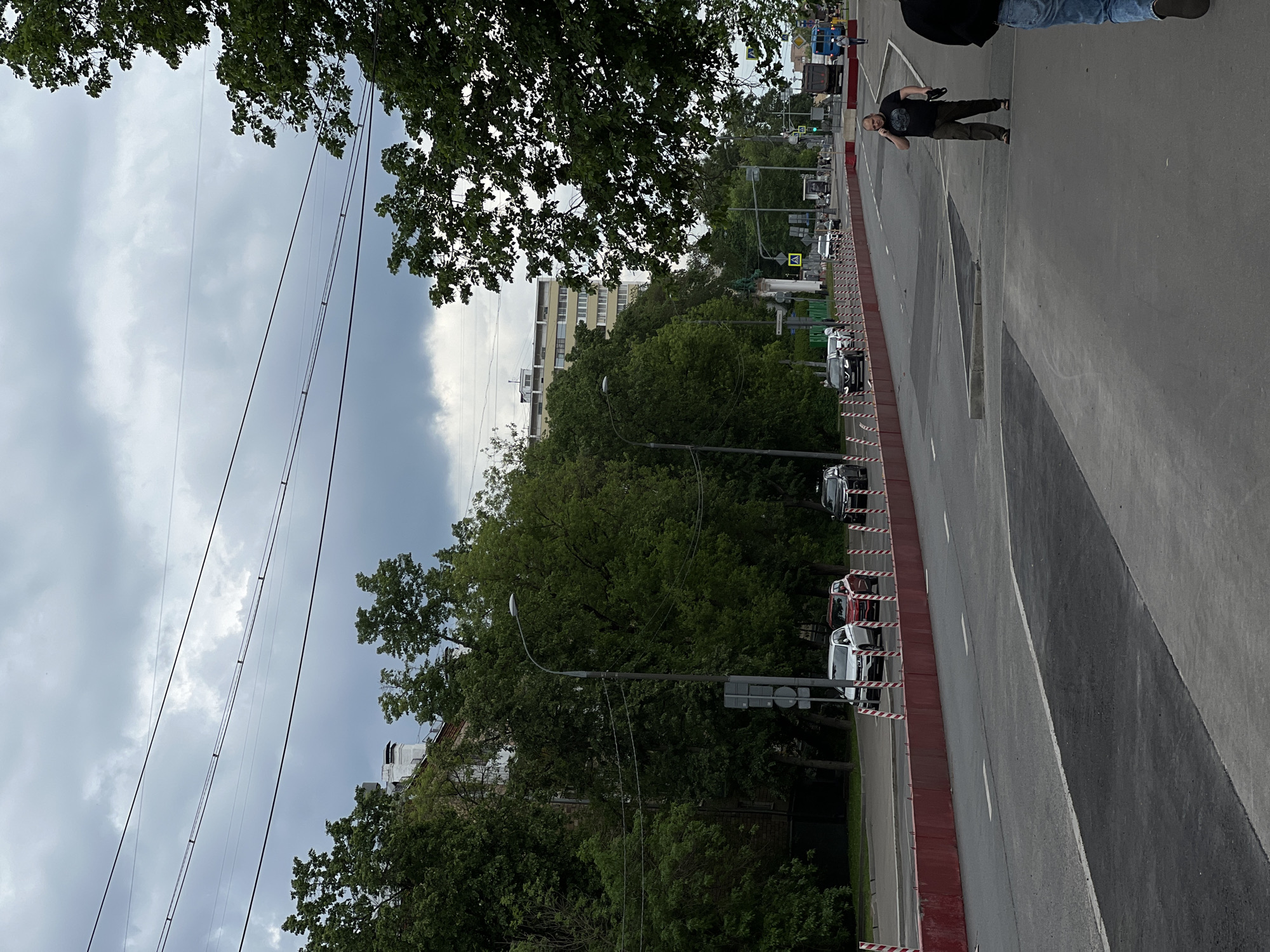 The image shows a street scene in Moscow, Russia. The view is along a multi-lane road with cars parked along the curb. Trees line the road, obscuring buildings. A man walks on the sidewalk in the upper right corner of the frame. Overhead wires crisscross the sky. The sky is overcast. A red and white barrier runs along the curb. Traffic lights are visible in the distance.