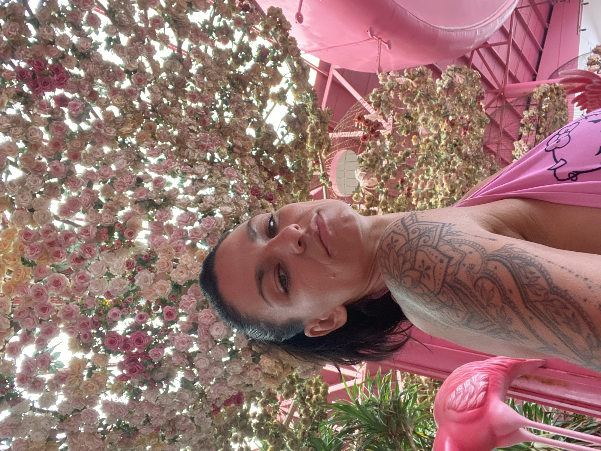 This image, taken in Tien Thanh Ward, Vietnam, features a woman captured in a low-angle shot, looking towards the camera with a subtle smile. Her head is tilted, and she is seen from the upper torso. She has dark hair and an extensive, intricate tattoo covering her right arm and shoulder with patterns resembling Mandalas and natural elements. She is wearing a bright pink top, part of which shows a black outline of a cartoon character.
The scene is highly stylized and vibrant, dominated by shades of pink. Above and around the woman, a dense canopy of artificial roses in various hues of pink, white, and peach creates a lush, floral ceiling. Bright daylight filters through these flowers, suggesting the photo was taken during the day.
In the background and to the right, a large architectural structure, including beams and support elements, is entirely painted in a bright pink color, reinforcing the themed environment. A decorative pink flamingo statue is visible in the bottom right corner, partially obscured by green foliage, further enhancing the whimsical atmosphere. The overall setting suggests a uniquely decorated indoor or semi-outdoor space, possibly a themed cafe or restaurant. No visible text is identifiable in the image.
