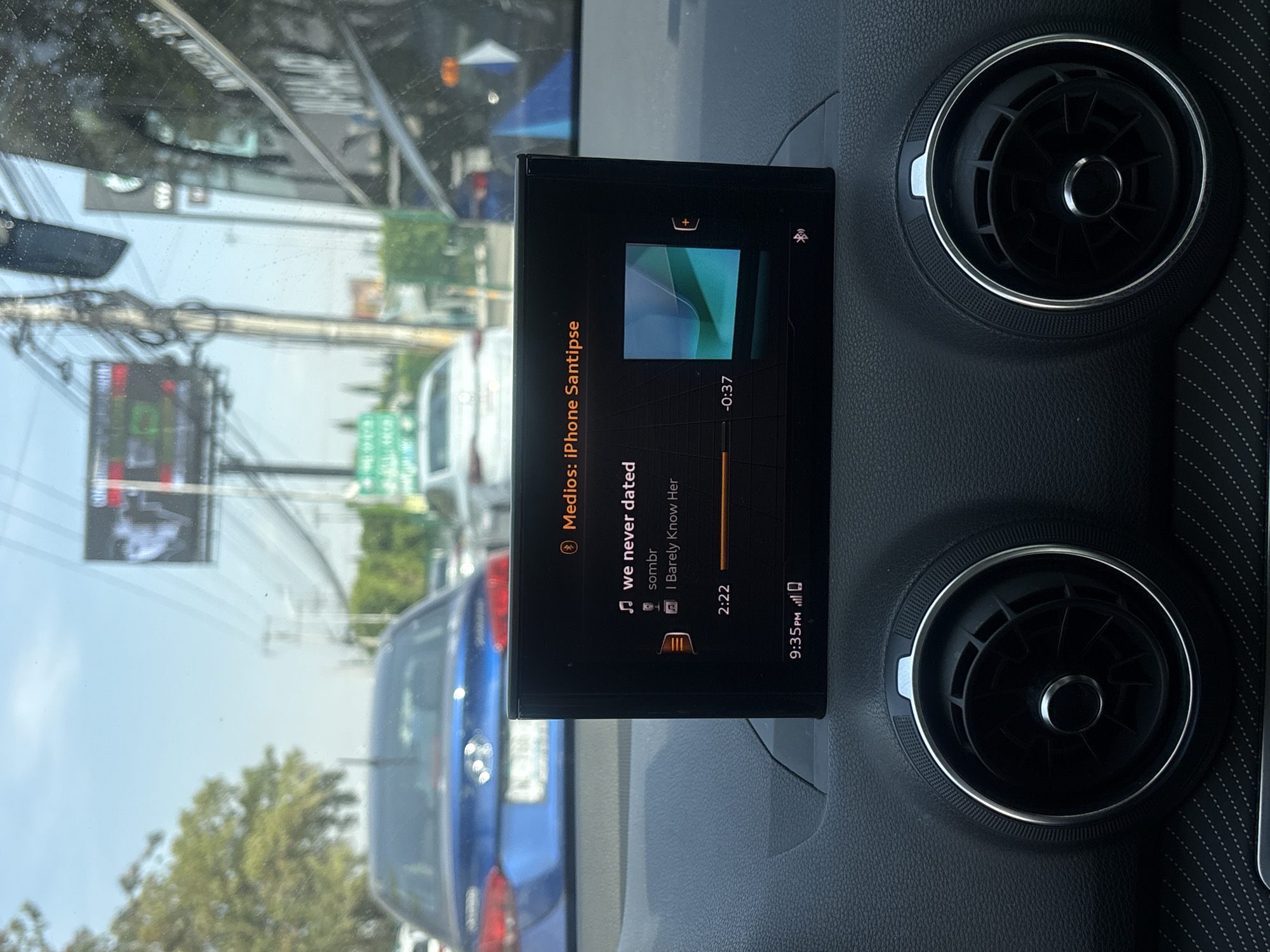 The image displays the interior of a car, focusing on its dashboard and an active infotainment screen, with reflections of an outdoor street scene on the windshield. On the right, two circular air vents, characteristic of an Audi, feature metallic bezels and a textured surrounding. The car's dashboard is covered in a dark material with subtle stitching visible on the far right.
The infotainment screen, positioned centrally, is displaying music playback information from a device labeled "Medios: iPhone Santipse." The currently playing song is titled "we never dated" by "sombr," from the album "I Barely Know Her." The track's current timestamp is 2:22, with -0:37 remaining. The screen also shows a digital clock indicating "9:35 PM," though the bright, sunny conditions visible through the windshield suggest it is daytime. Other icons for battery level and wireless connectivity are present.
Through the windshield, a busy urban environment in Puebla, Mexico, is reflected and visible. Numerous overhead power lines are prominent, along with blurred buildings, green trees, and other vehicles, including a blue sedan and a white van. A traffic light showing both red and green elements is partially visible. The scene suggests the car is either stopped or moving slowly in traffic on a clear, sunny day. No people are explicitly visible in the frame.