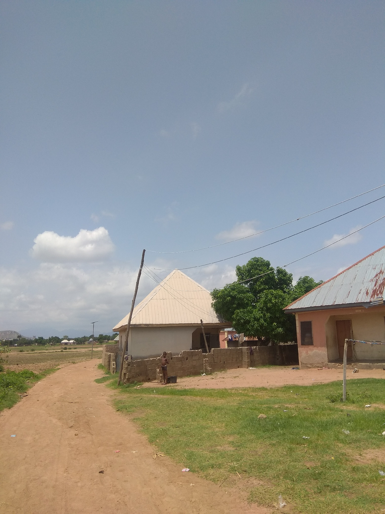 A dirt road leads through a rural village in Jalingo, Nigeria. The sky is a clear blue with scattered white clouds, suggesting it is a bright, sunny day. Two simple houses are visible, one with a light-colored, peaked metal roof and the other with a weathered, corrugated metal roof. A wall constructed of rough stones surrounds the first house. A young boy stands near the wall of the first house, appearing to be engaged in some activity, possibly washing or preparing something in a bucket. A tall wooden pole with electrical wires runs near the first house. A large, leafy green tree stands between the two houses. The ground is a mixture of dirt and sparse green grass, with some litter scattered about. In the distance, a few more buildings and trees can be seen, with a hazy mountain range in the far background. The scene appears to be a typical day in a Nigerian village, with a focus on the everyday life and structures of the community.