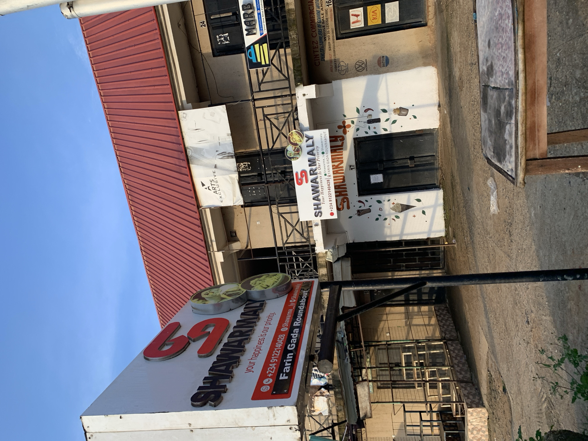 The image shows the exterior of a commercial building with multiple businesses. A prominent sign for "SHAWARMALAY" with a phone number and address, "Farin Gada Roundabout", is visible. Below the sign, the same business name is painted on a white wall, adorned with illustrations of food items. Adjacent to Shawarmaly, there are other businesses with signage such as "MARB" and "CINTEZ COMMERCIAL". The building has a red metal roof and a balcony. The foreground shows a concrete ground and a rustic wooden table with a worn surface. The overall setting appears to be in an urban area, likely in Rafin Par, Nigeria, based on the mention of "Farin Gada Roundabout" which is a known location in Jos, Plateau State, Nigeria. The sky is clear and blue.