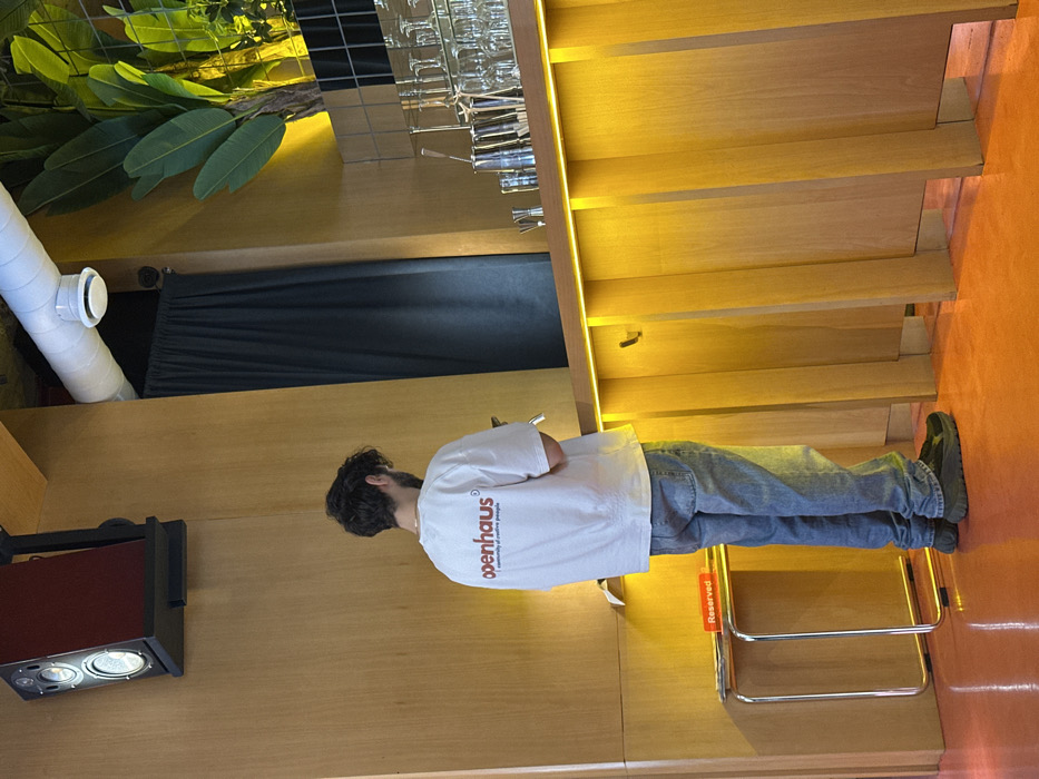 An interior shot depicts a male individual standing with his back to the viewer within a modern establishment. The location is identified as Openhaus, a creative space at Aram St 75, Yerevan, Armenia, based on the logo visible on the subject's shirt. The man has dark, curly hair and a beard, and is wearing a white t-shirt with "openhaus" in red and "a community of creative people" below it, light blue jeans, and dark, platform-soled sneakers. He is holding and looking down at a smartphone. The environment features a bright orange floor and light-colored wooden wall paneling. To the man's right is a wooden bar counter with a horizontal yellow LED strip light, above which multiple inverted clear glasses and metal bar tools are arranged. Behind the bar, dark tiled sections and green leafy plants are visible. Above, a white ventilation pipe runs along the ceiling with a circular vent. To the man's left, a "Reserved" sign in red sits on a metallic, minimalist stand. A dark curtain is visible in the background. A two-driver speaker in a dark enclosure is mounted on a wall bracket in the upper left.