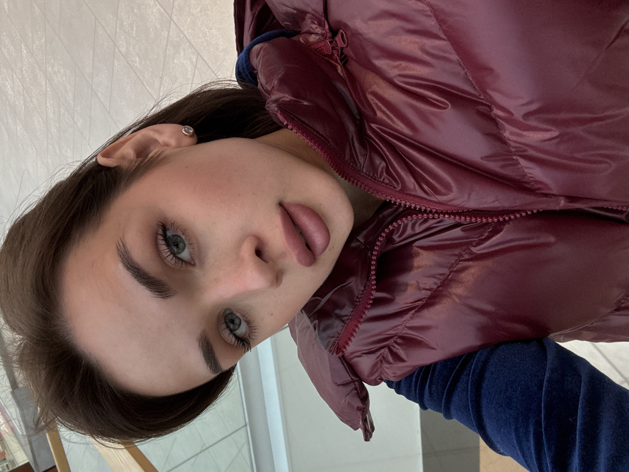 A young woman with fair skin, light eyes, and dark eyebrows is posing for a picture indoors. She is wearing a burgundy puffer vest over a dark blue velvet-like top. She is wearing a small stud earring. The background appears to be a tiled floor and a wall. The location is Perm, Russia.