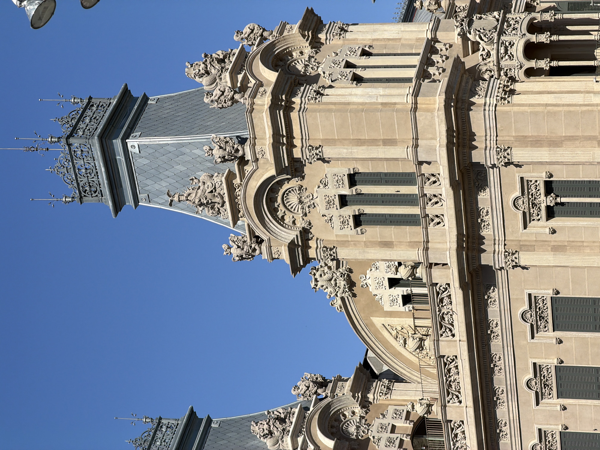 The image shows a close-up of the upper facade of a building in Barcelona, Spain, featuring intricate architectural details. The building is constructed from light-colored stone and exhibits a style characterized by ornate sculptural elements, arched windows with dark shutters, and decorative cornices. Several turrets or decorative spires crown the building, topped with filigree metalwork. The clear blue sky serves as the backdrop. The building's location is the **Port of Barcelona**, specifically the **Edifici de la Llotja de Mar** (Maritime Stock Exchange Building). No people or specific actions are visible in the image. The overall impression is one of historical grandeur and detailed craftsmanship.