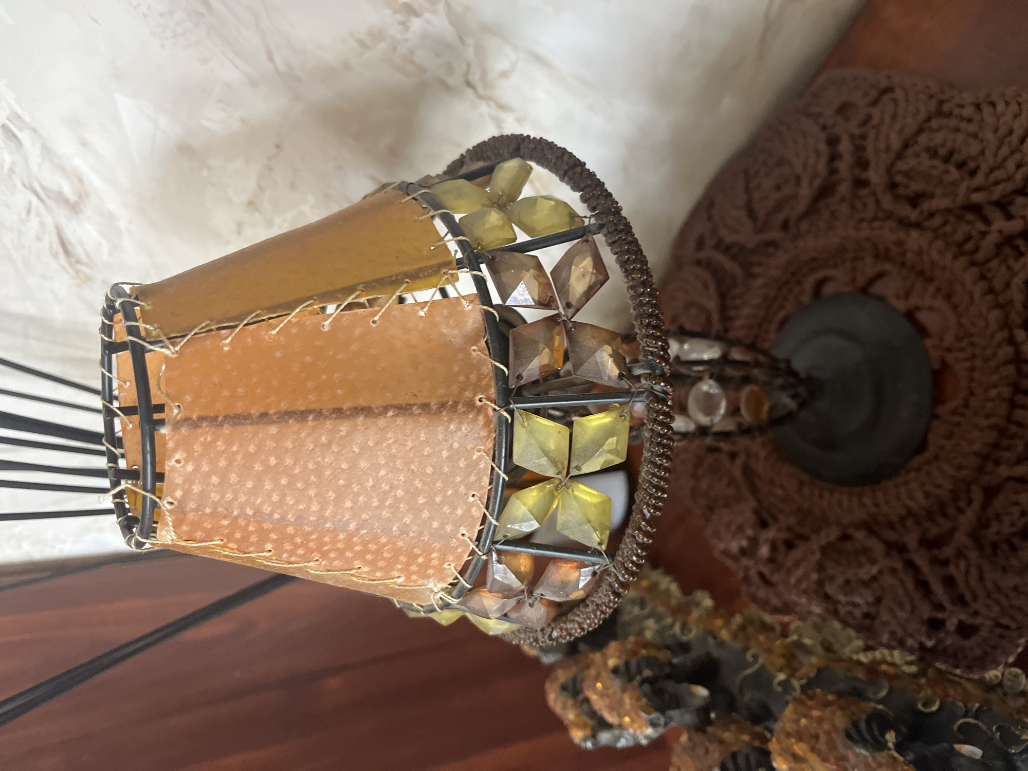 This is a medium shot of a decorative lamp with a conical shade made of overlapping pieces of amber-colored material, stitched together with thin, light-colored string. The shade is supported by a black metal frame. Dangling from the bottom of the shade is a decorative fringe made of square, amber-colored glass beads. The lamp is situated on a wooden surface, possibly a table. To the left of the lamp, a portion of another decorative item with similar amber-colored embellishments is visible. The background consists of a dark brown curtain on the left and a textured, light-colored wall on the right. There are no people or animals present in the image. The scene appears to be an indoor setting.