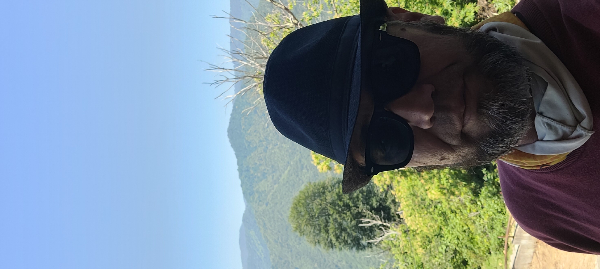 The image shows a close-up of a person wearing a dark blue hat and sunglasses, with a scenic mountainous landscape in the background. The person has a grey beard and is wearing a maroon sweater over a patterned neck gaiter. The setting appears to be outdoors, with lush green trees and hills under a clear blue sky, suggesting a sunny day. The angle of the photograph is such that the person is in the foreground, with their face turned partially away from the camera, looking towards the right of the frame. The background shows rolling hills covered in dense vegetation, indicative of a natural, possibly rural or mountainous, environment. The lighting suggests it is daytime, likely morning or afternoon, given the clear skies and bright illumination of the landscape. There are no visible text elements in the image.