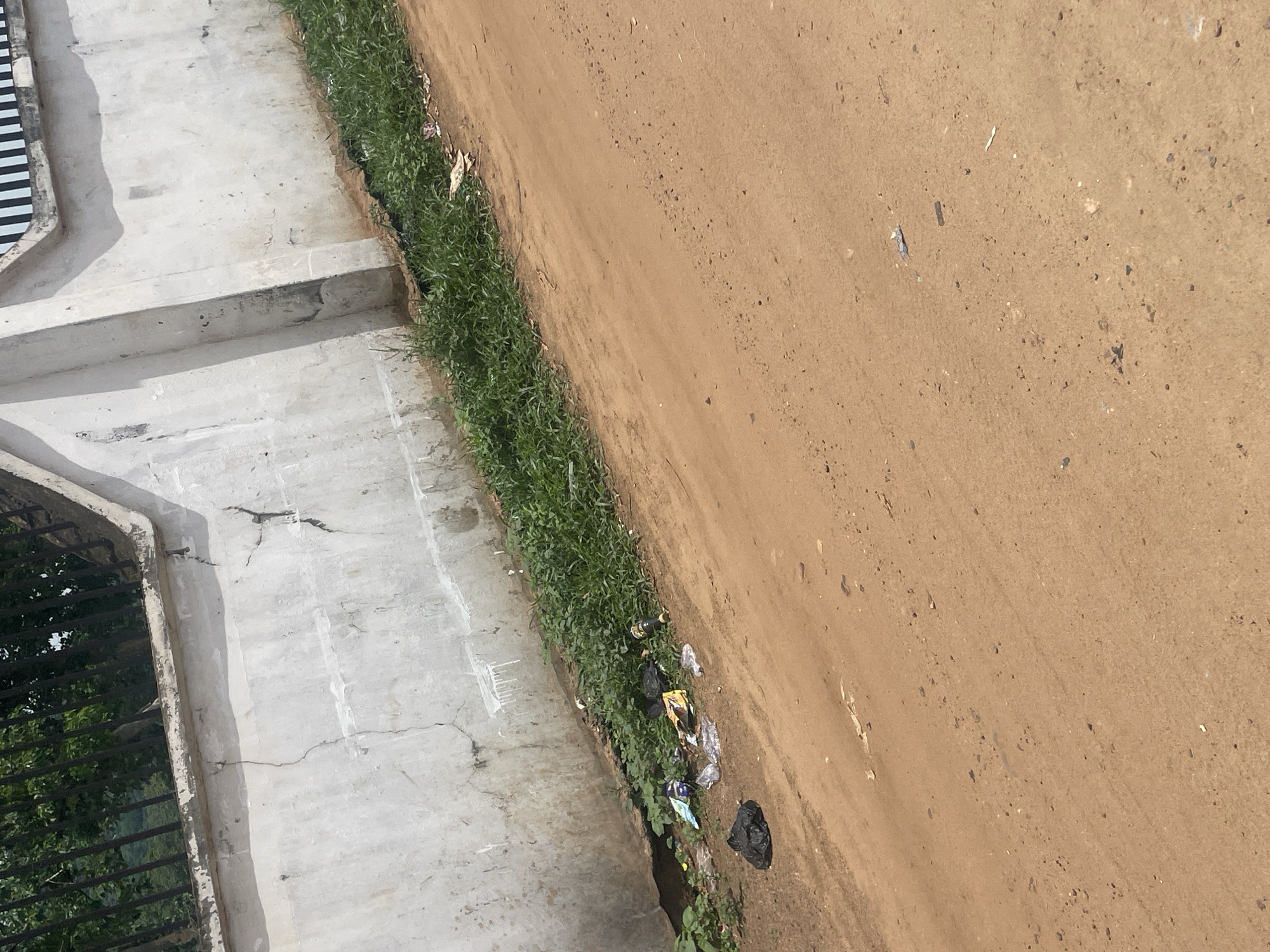 The image shows a close-up, high-angle view of a dirt path next to a concrete ledge and some overgrown grass. The dirt path, which takes up the majority of the frame, is a light brown color with small pebbles and debris scattered across it. The concrete ledge runs along the left side of the image, appearing worn and cracked in places. To the left of the concrete ledge, there is a dark metal grate that partially obscures a view of green foliage and trees, suggesting an outdoor setting.
Along the edge where the concrete meets the grass, there is a noticeable amount of litter. This includes plastic bags, wrappers, and what appears to be a bottle. The grass itself is green and somewhat unkempt, growing in a dense strip between the concrete and the dirt.
The lighting suggests it is daytime, likely with bright sunlight given the shadows and the clear visibility. There are no people or animals visible in the frame. The scene depicts an outdoor area, possibly an unpaved walkway or the edge of a construction site, in a location with moderate vegetation. The presence of litter indicates a lack of regular maintenance or public waste disposal. There is no discernible text in the image. The overall impression is one of an unkempt outdoor environment.
