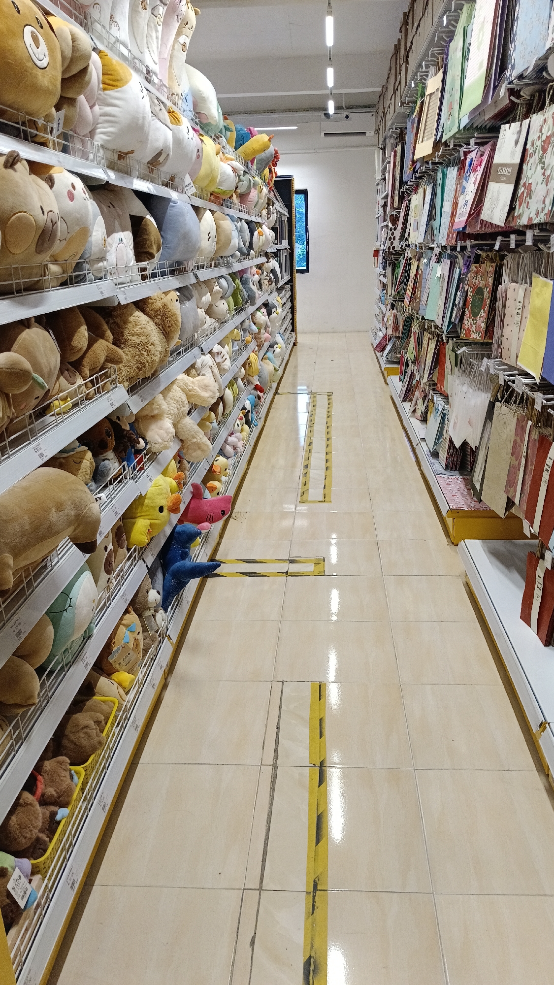 The image shows an aisle in a retail store, likely a gift shop or a toy store, in Gunting Saga, Indonesia. The aisle is flanked by tall shelves filled with merchandise. On the left, shelves are stacked with an abundance of plush toys in various sizes and colors, including teddy bears, cats, and animal-shaped pillows. On the right, shelves display an assortment of gift bags and stationery items, with patterns of flowers and other decorative designs. The floor is tiled in a beige color, with yellow and black striped floor markings indicating social distancing guidelines. Fluorescent lighting fixtures are visible on the ceiling. There are no people visible in the image. The store appears to be well-stocked and organized, with a focus on soft toys and gift-related items.
