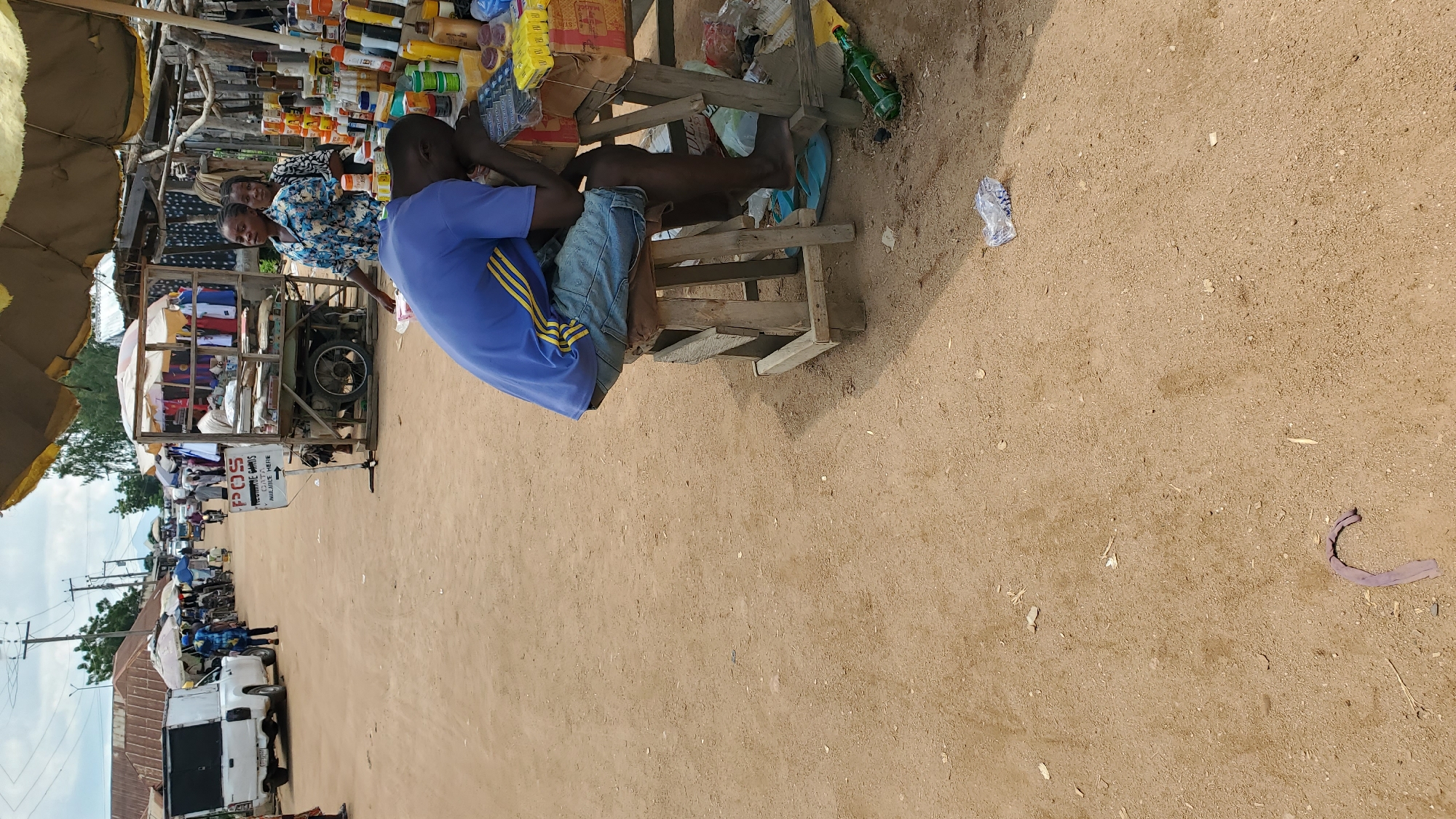 This is a medium shot, outdoors, in what appears to be a market street in Jalingo, Nigeria. The ground is dirt with scattered debris. In the foreground, a young Black man wearing a blue t-shirt with yellow stripes and denim shorts sits slumped forward on a wooden stool, his head resting on a table. The table is laden with various goods, including what appear to be toiletries and boxed items. To the right of the man, a green glass bottle lies on the ground, along with some crumpled plastic. In the background, the market continues with stalls selling clothes, and people are walking and riding bicycles. Two Black women stand behind the table, looking towards the left of the frame. A white pickup truck is parked on the left side of the road. A sign on a cart reads "POS Recharge Cards Available Here". The sky is overcast.