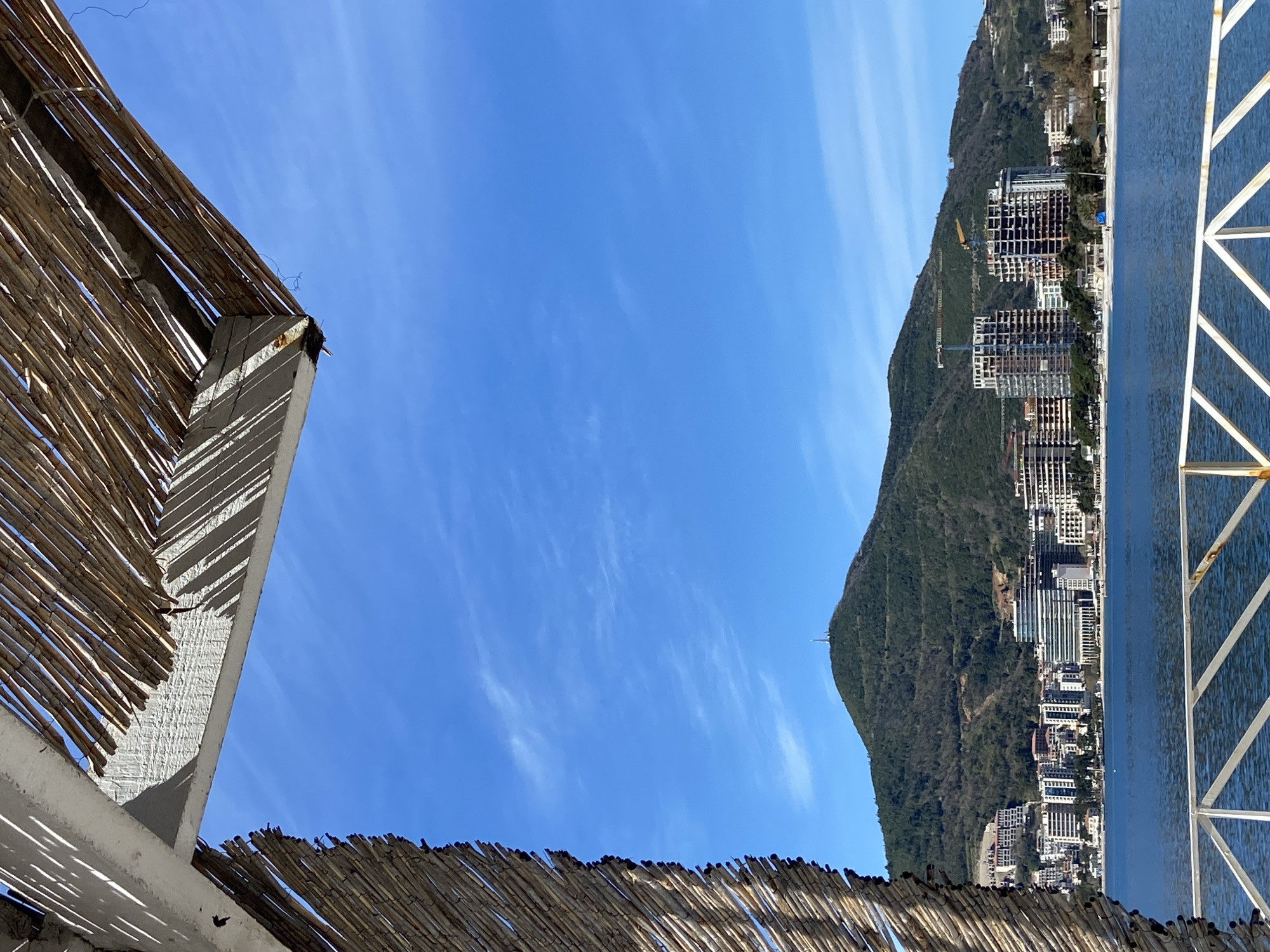 The image presents a view of Budva, Montenegro, from beneath a thatched structure, likely a beachside shelter. The sky is a vibrant blue with wispy clouds. To the right, a mountain covered in dense vegetation forms a backdrop for the city, which is densely built with high-rise buildings along the coastline. A portion of a pool or a body of water with a white, geometric structure is visible at the far right edge. The perspective is tilted.