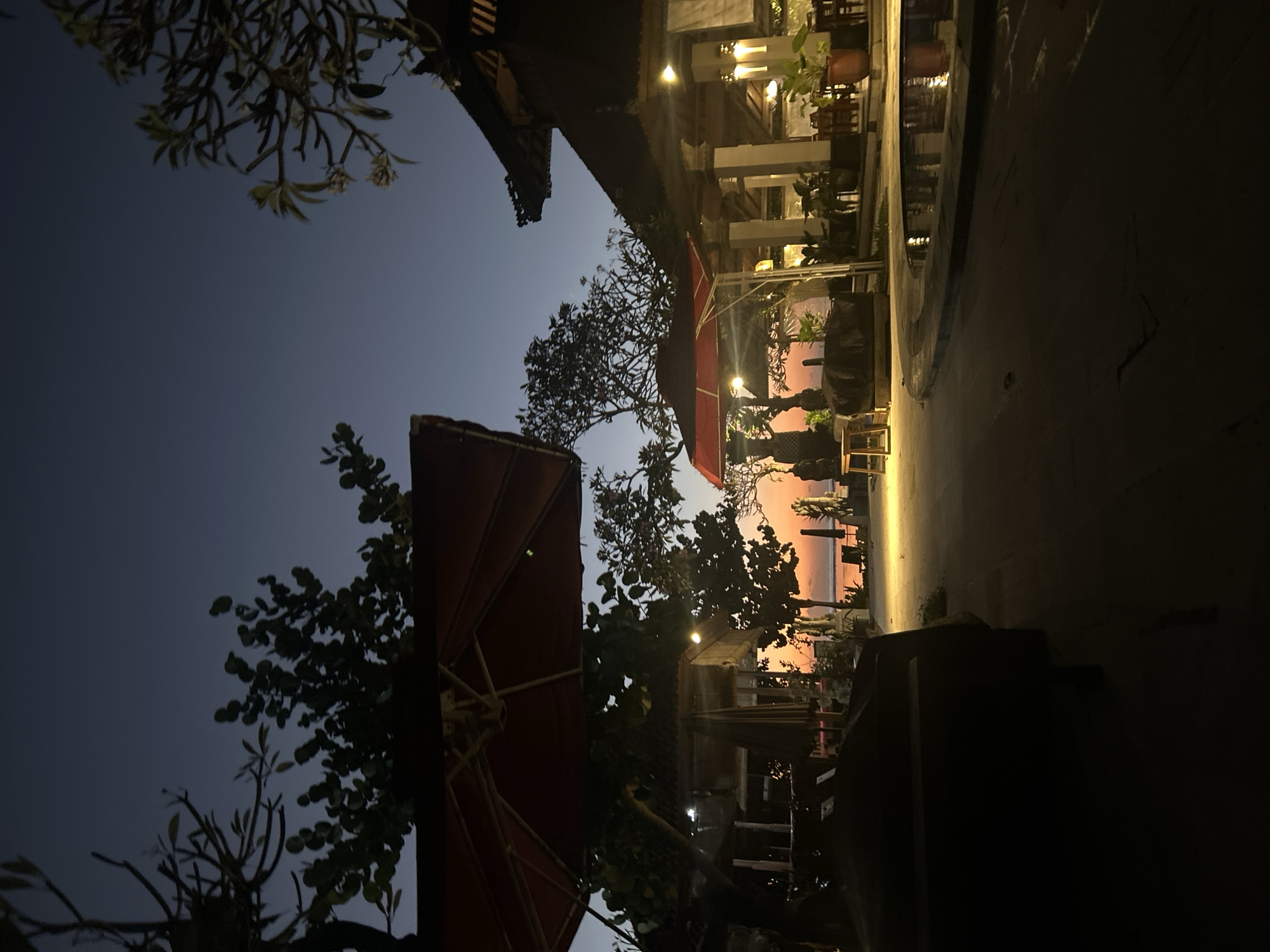 The image depicts a dimly lit outdoor scene at dusk, likely a resort or restaurant area by the beach. In the foreground, a large dark object, possibly a lounge chair or a seating area, is silhouetted against the brighter background. To the left, a red umbrella provides shade. Further back, there is a building with a traditional pitched roof, illuminated by soft lights, and several potted plants. A small body of water, perhaps a pool, is visible to the right, reflecting the lights. The background shows a beach with chairs and umbrellas, and the ocean with the sky transitioning from the warm hues of sunset to a deep blue. Trees with sparse foliage are scattered throughout the scene. The overall atmosphere is serene and peaceful. This location is likely in the Sanur area of Denpasar, Bali, Indonesia, given the architecture and the beachfront setting.