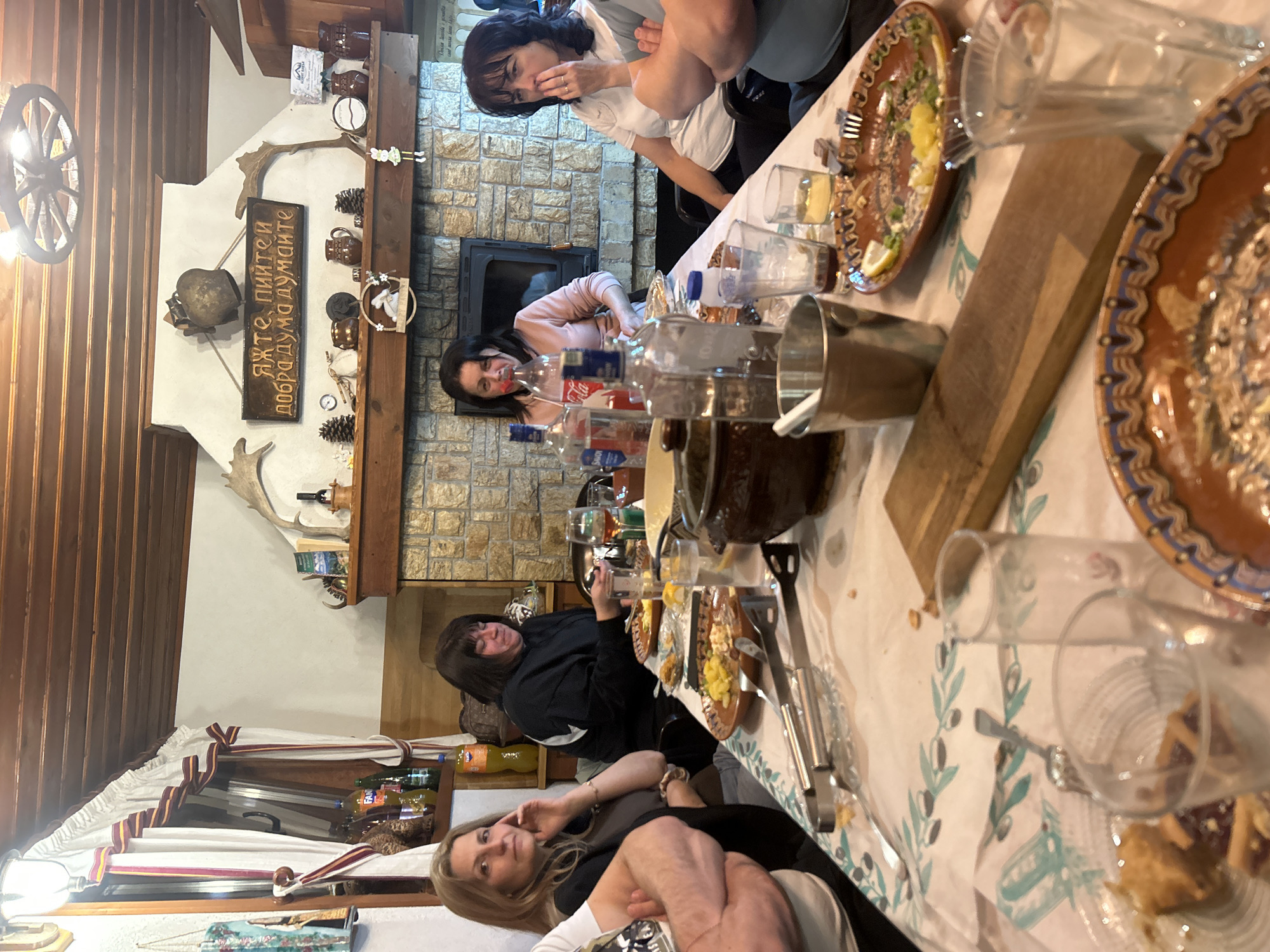 An indoor scene in what appears to be a traditional Bulgarian restaurant or guesthouse in Zlatograd, Bulgaria, depicts a social gathering of approximately five to six women around a long wooden table. The room features rustic decor with wooden ceiling beams, a stone fireplace with a black metal insert, and shelves adorned with traditional pottery, antlers, and pinecones. Artificial lighting, including a prominent wagon-wheel-style chandelier, illuminates the space, suggesting it is evening or nighttime.
The women, dressed casually, are seated around the table, which is covered with a white tablecloth featuring an olive branch pattern. The table is laden with partially consumed food, including salads with lemon, and various drinks in glasses and bottles (e.g., Coca-Cola, clear spirits). Traditional brown and orange ceramic plates with intricate patterns hold food and desserts. A large earthenware pot and a metal bucket are also on the table. Several women are looking towards the camera, some smiling, indicating a friendly and relaxed atmosphere.
A notable decorative element is a wooden plaque mounted above the fireplace, bearing the Cyrillic text: "ЯЖТЕ, ПИЙТЕ И ДОБРА ДУМА ДУМАЙТЕ", which translates to "Eat, drink and speak good words." This phrase is a common Bulgarian saying, reinforcing the cultural context. The overall impression is one of a convivial meal or celebration among friends or family in a cozy, traditionally decorated setting.