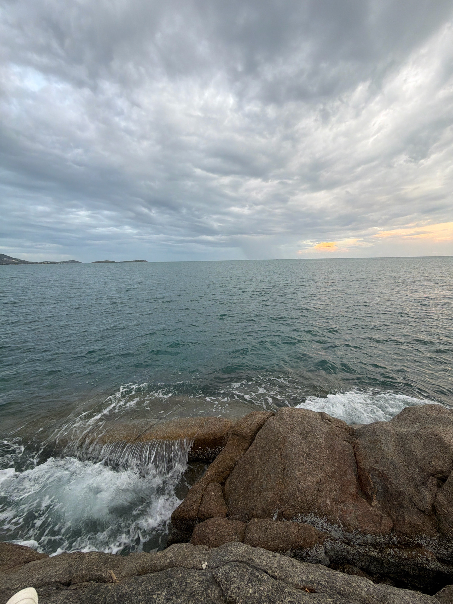 The image depicts a coastal scene at dusk in the Koh Samui District of Thailand. The sky is overcast with a thick layer of gray clouds, but a faint golden glow is visible on the horizon, suggesting the sun is setting. The sea is calm, with a moderate swell and small waves lapping against a rocky shoreline in the foreground. The rocks are of various sizes and colors, ranging from dark gray to reddish-brown. A small cascade of water flows over a flat rock into the sea. In the distance, a few islands are visible on the horizon. There are no people or animals present in the image. The overall impression is one of a serene and somewhat moody coastal landscape.