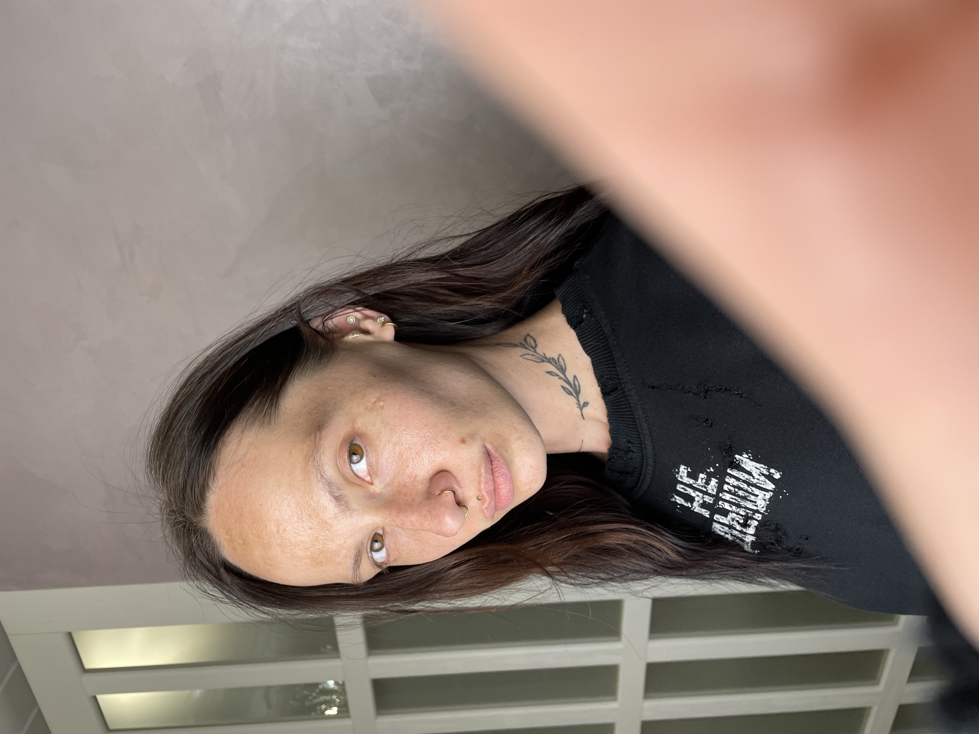 A young adult female with light complexion, brown hair, and hazel eyes is seen from a low-angle perspective. She has facial piercings in her left nostril and above her lip, and a tattoo of leaves on her neck. She wears a distressed black garment with faded white lettering. The background consists of a light-colored wall and a set of white, paneled doors or windows. There is no indication of Izhevsk, Russia, in the image.